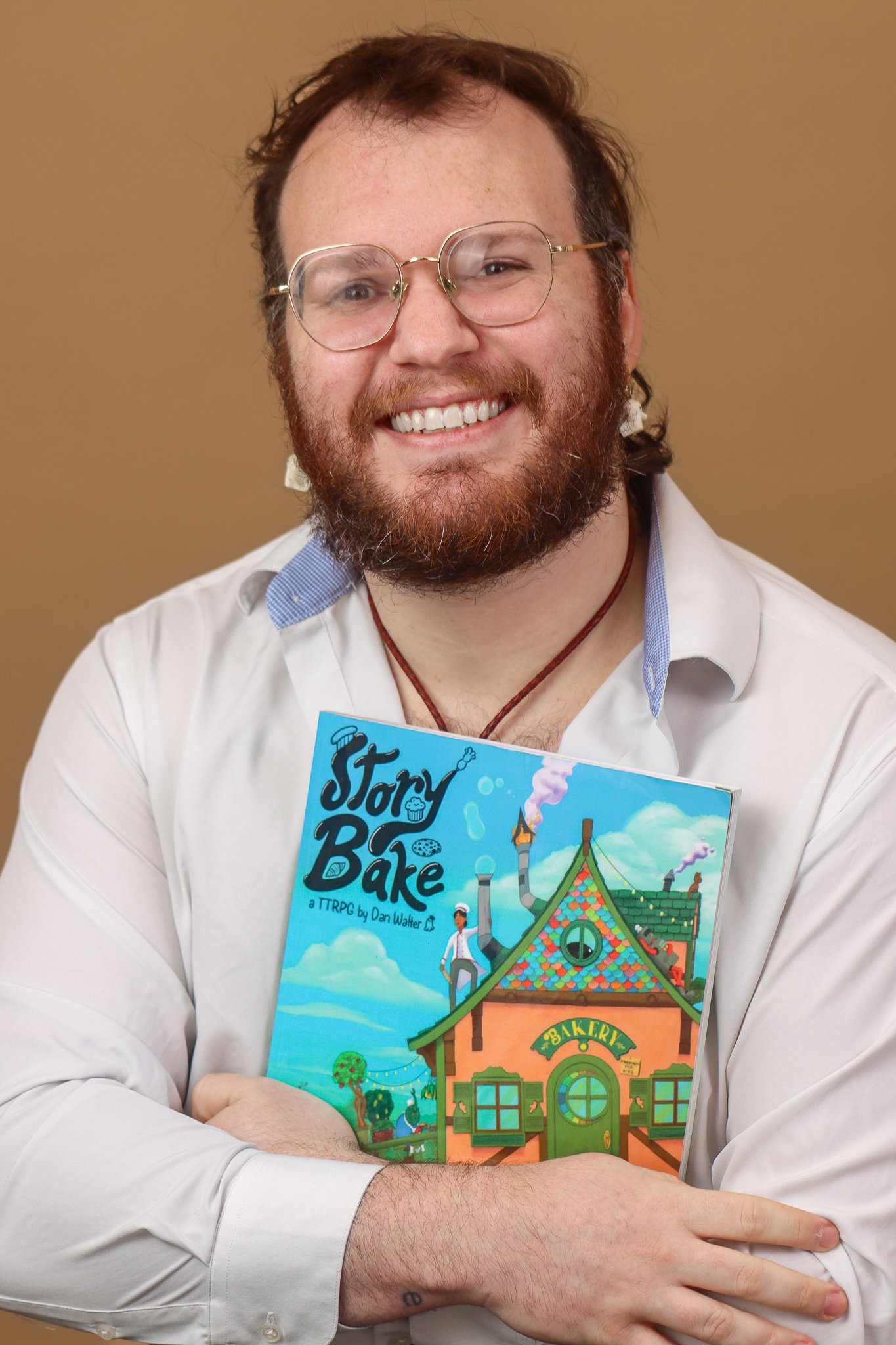 A man with glasses, a beard, and a smile wearing a white shirt, holding a colorful book titled "Story Bake," standing against a plain background.