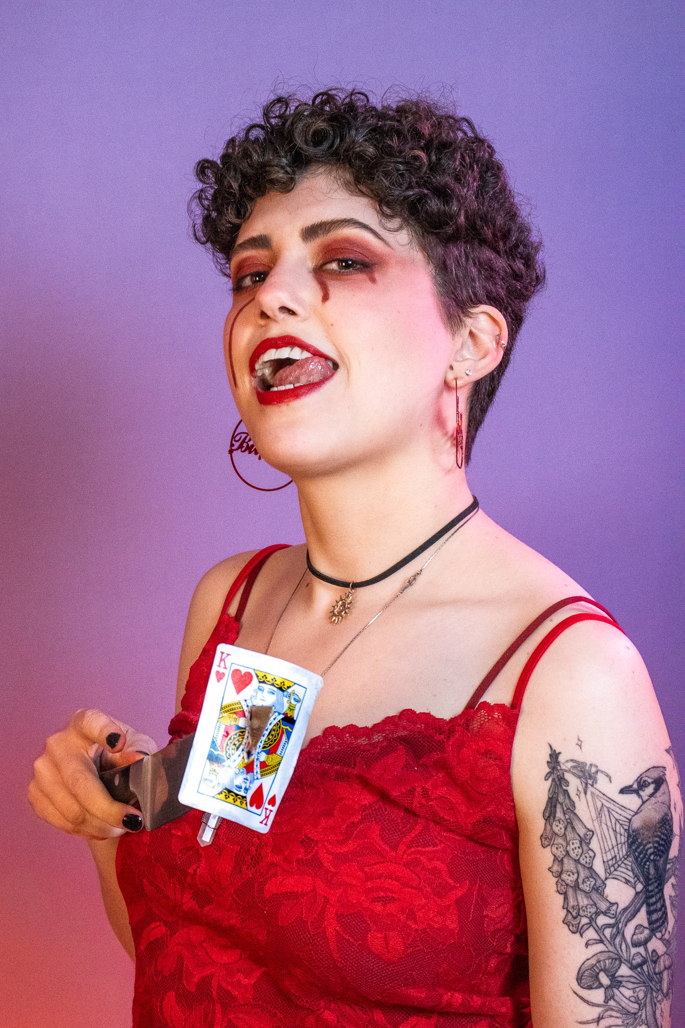A woman with short curly hair and tattoos on her arm, holding a playing card (King of Hearts) and a scalpel, dressed in red lace attire, with makeup featuring dark lipstick and eye makeup, posing against a purple background.