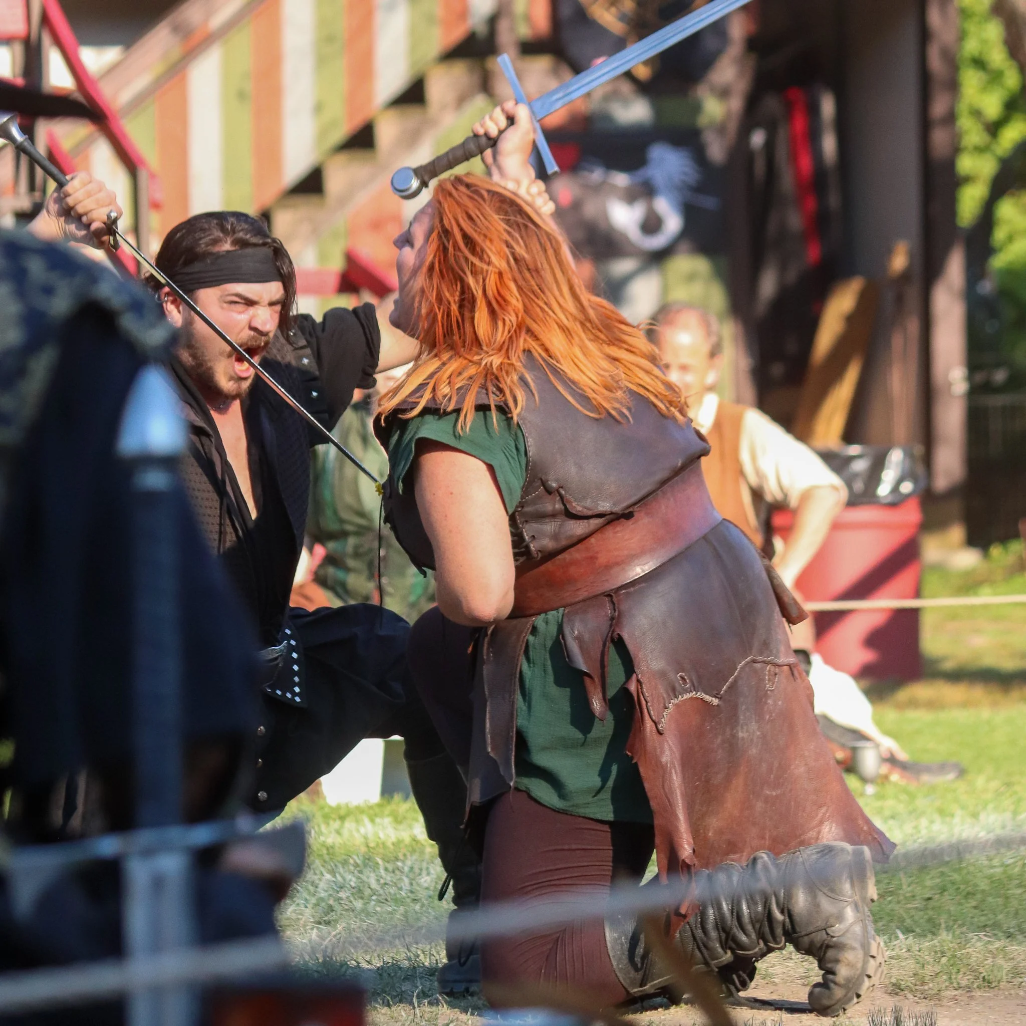 Two men engaged in a medieval combat reenactment or mock battle, brandishing swords and wearing period-appropriate costumes, with one man with red hair and leather attire.