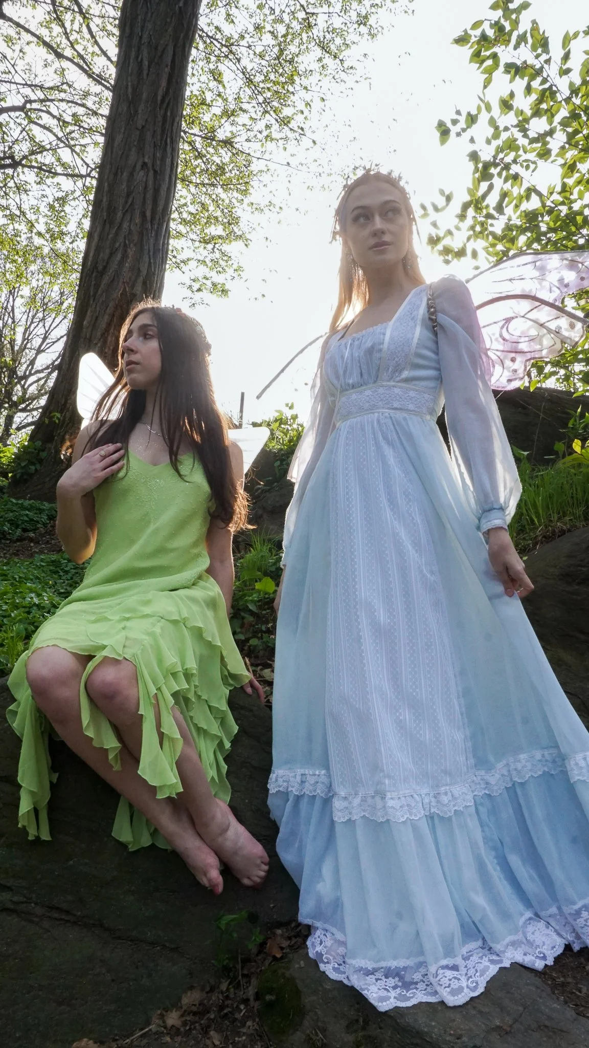 Two women with fairy wings in a forest, one sitting on a rock in a lime green dress, the other standing in a white dress with lace details. Sunlight filters through the trees.