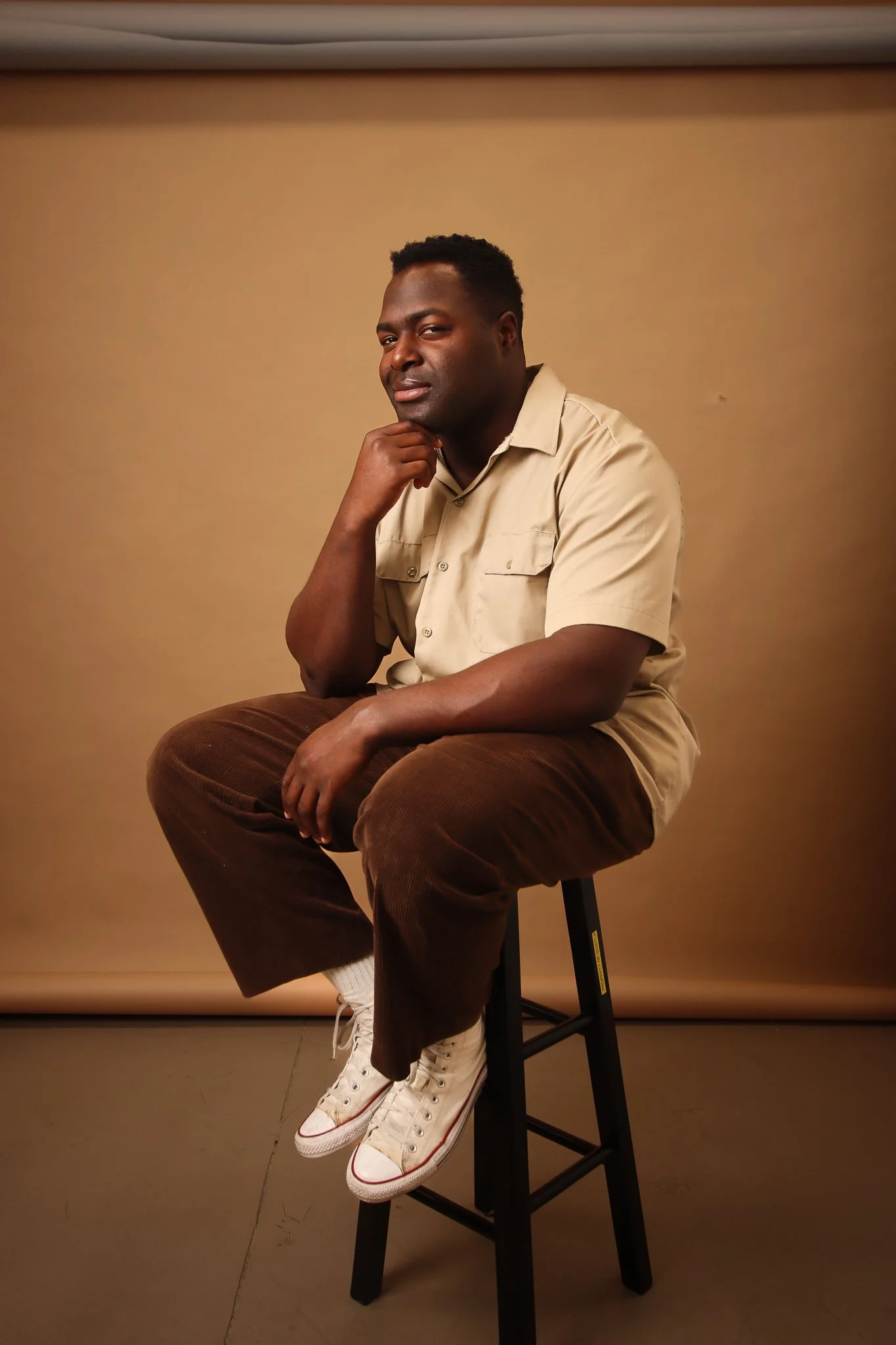 A man sitting on a black stool with his chin resting on his hand, wearing a beige short-sleeved shirt, brown corduroy pants, and white high-top sneakers, against a plain taupe background for a portrait photo.