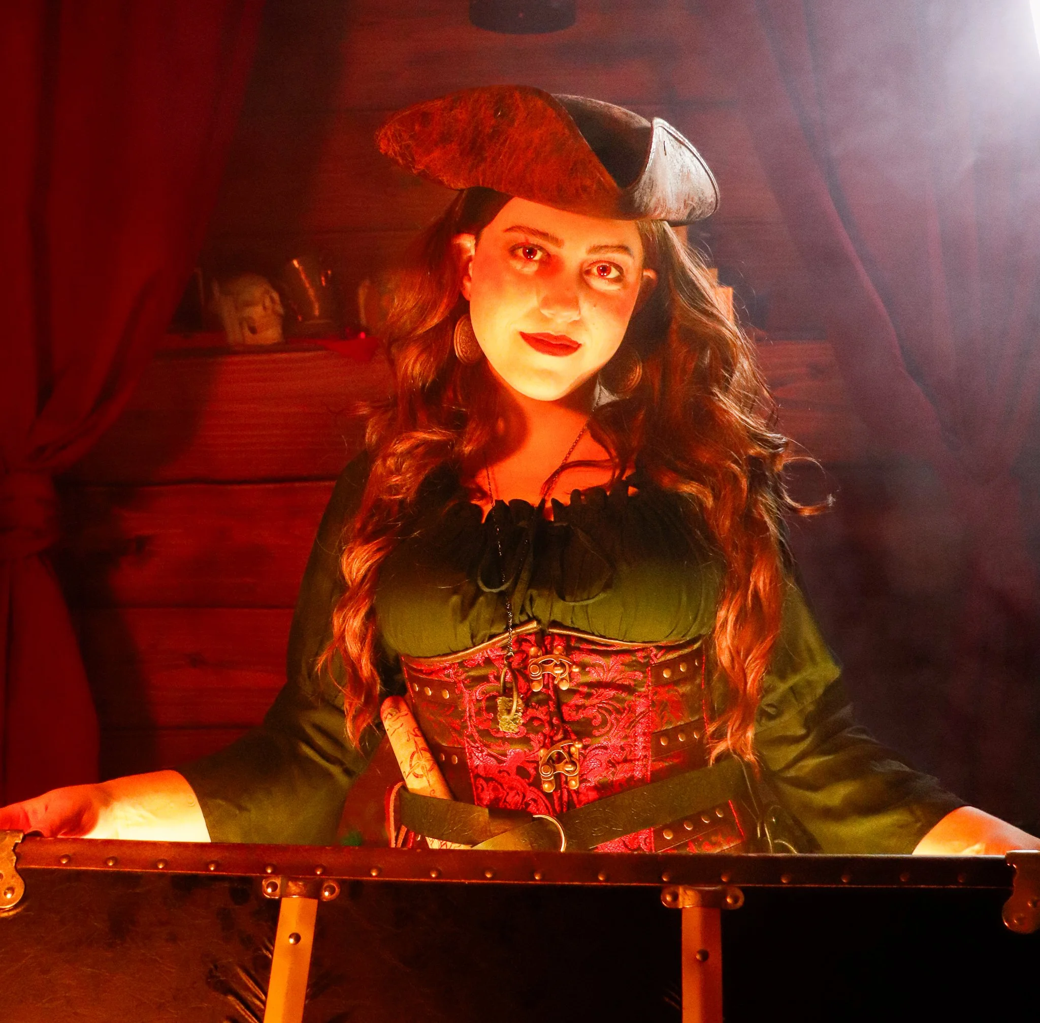 Woman with long curly hair wearing a historical costume with a decorative bodice and a tricorn hat, standing in a dimly lit room with red lighting.