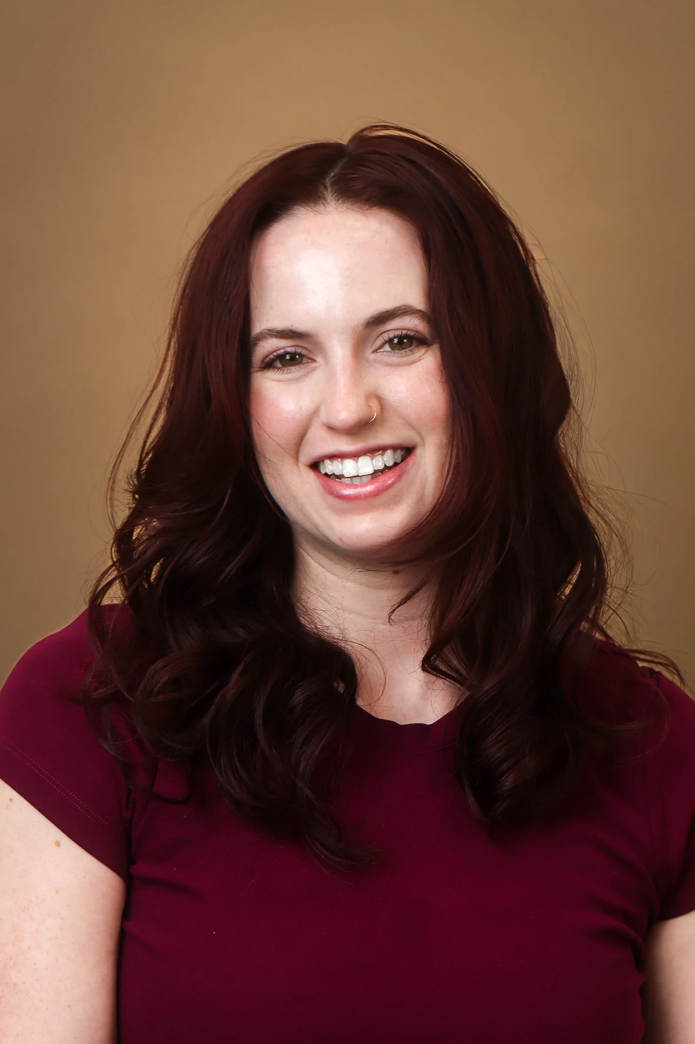 Close-up of a smiling woman with long, wavy red hair, wearing a maroon top, against a plain beige background.