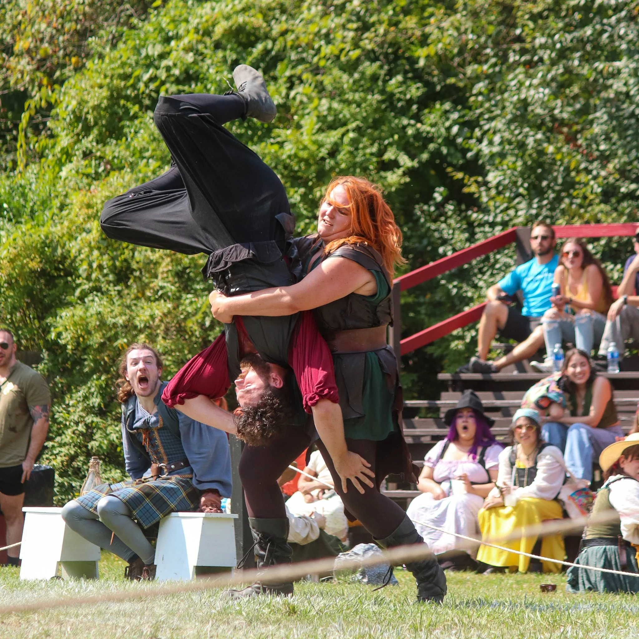 A woman with red hair and a black shirt is lifting a man with a black jacket and plaid pants onto her shoulders. They are outdoors with trees and people watching in the background.