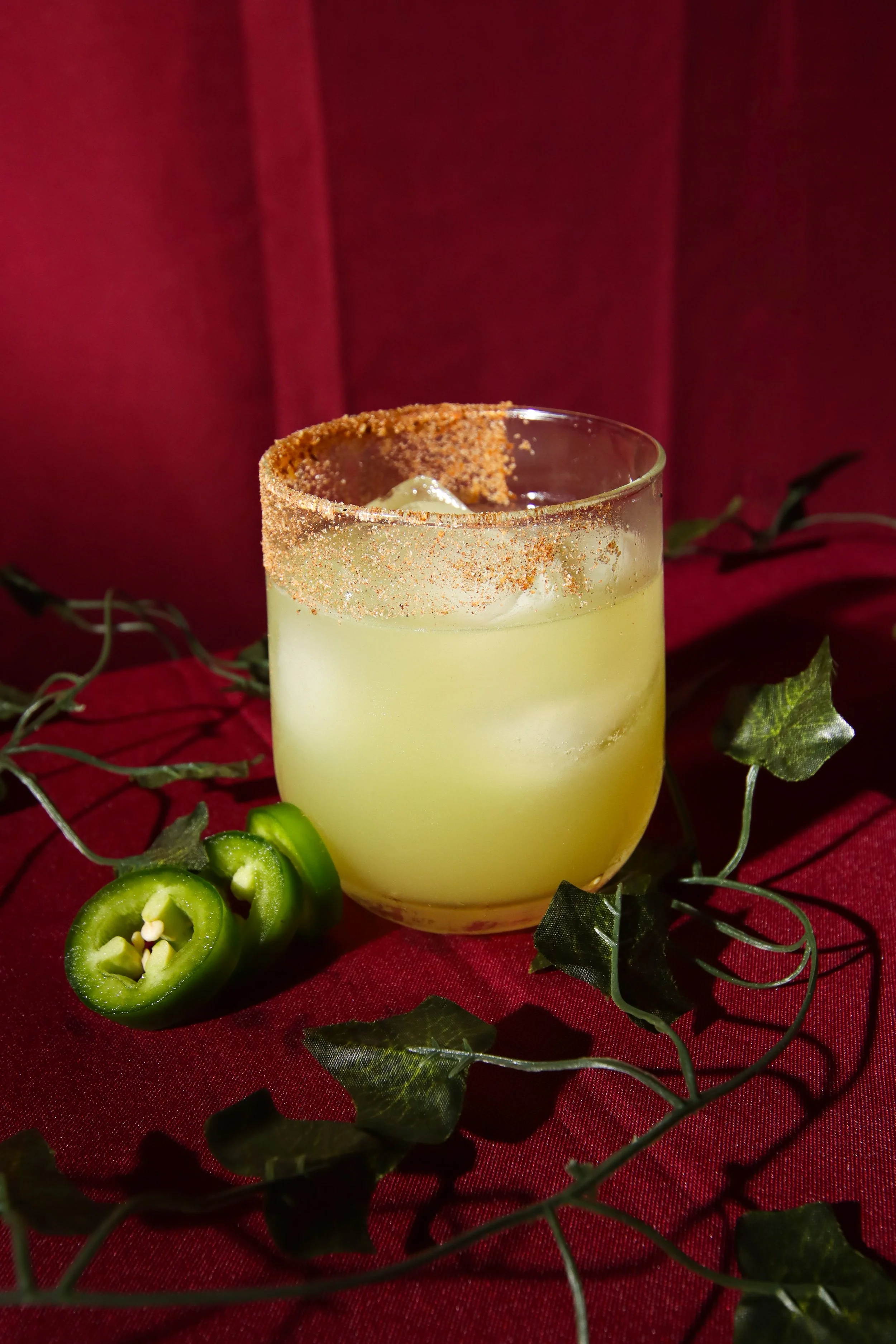 A cocktail glass with a greenish-yellow drink, garnished with a chili pepper rim and surrounding green chili slices, set on a red fabric background decorated with green leaves and vines.