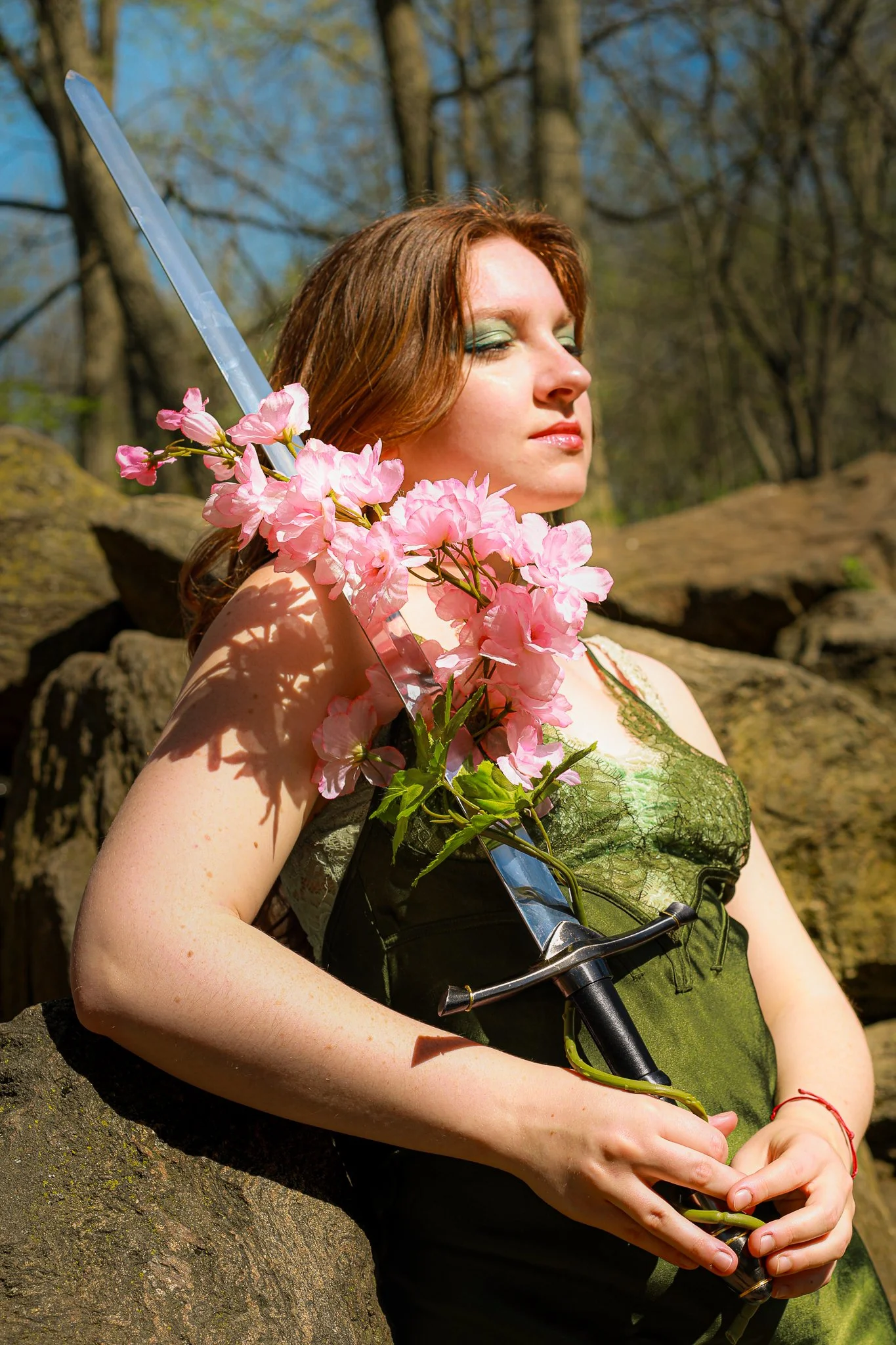 woman with closed eyes, holding a bouquet of pink flowers and a sword, outdoors with trees and rocks in the background