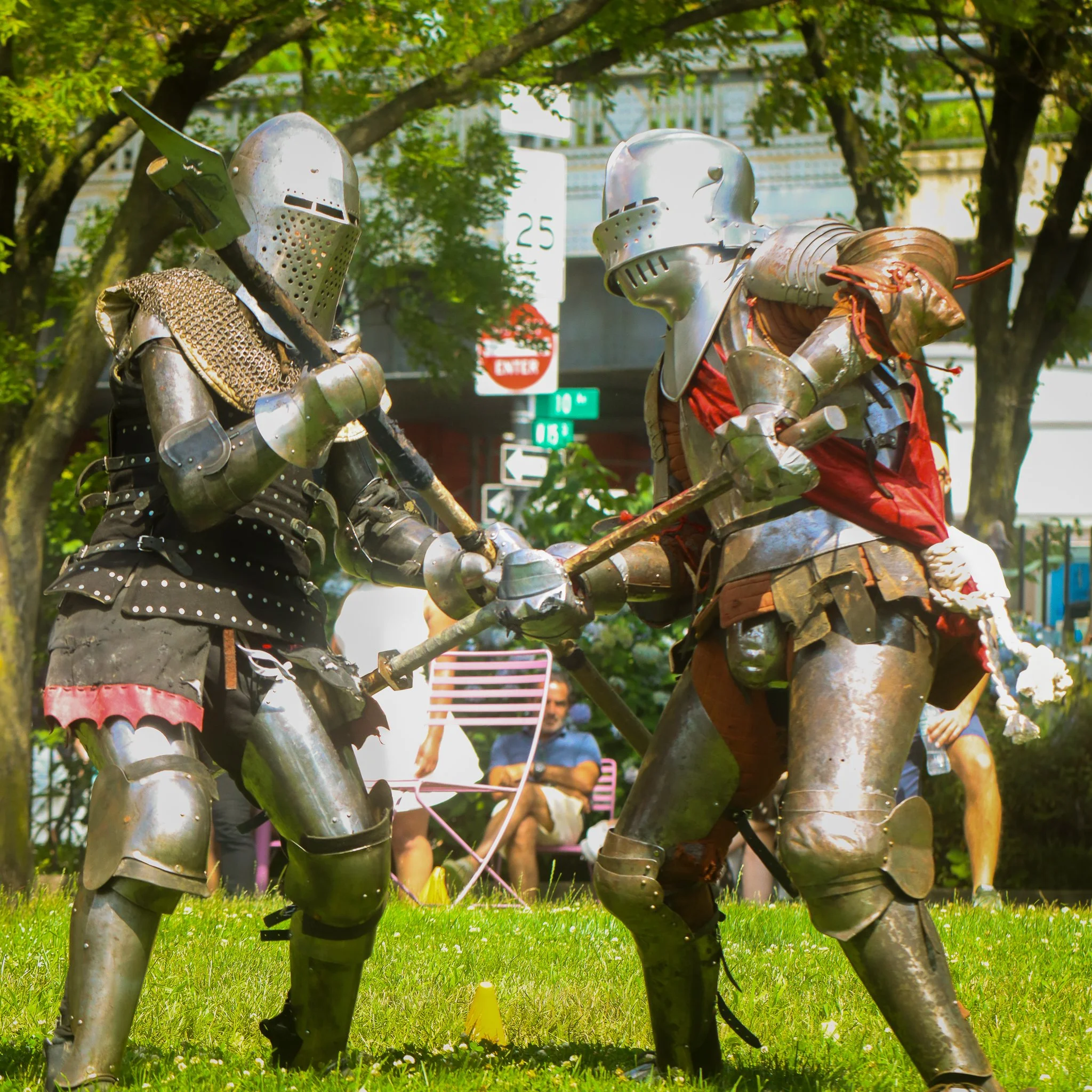 Two people dressed as medieval knights engaged in a staged combat or reenactment in a park, with green trees and street signs visible in the background.