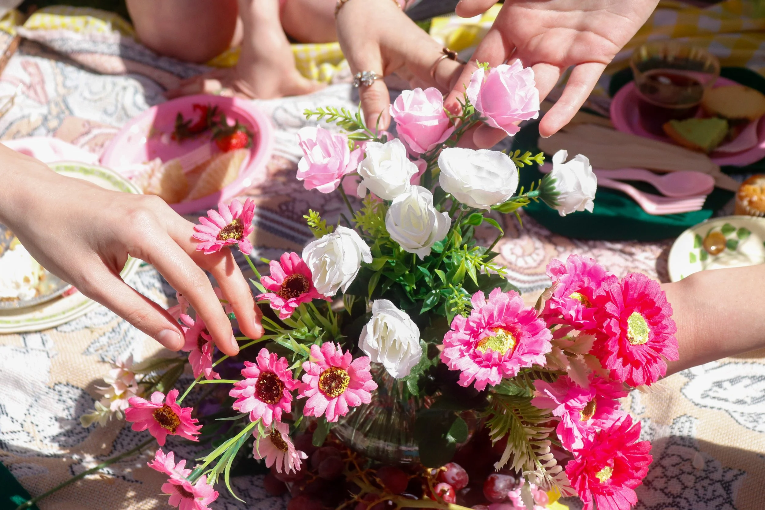 People arranging pink and white artificial flowers on a table with various plates of food and snacks.