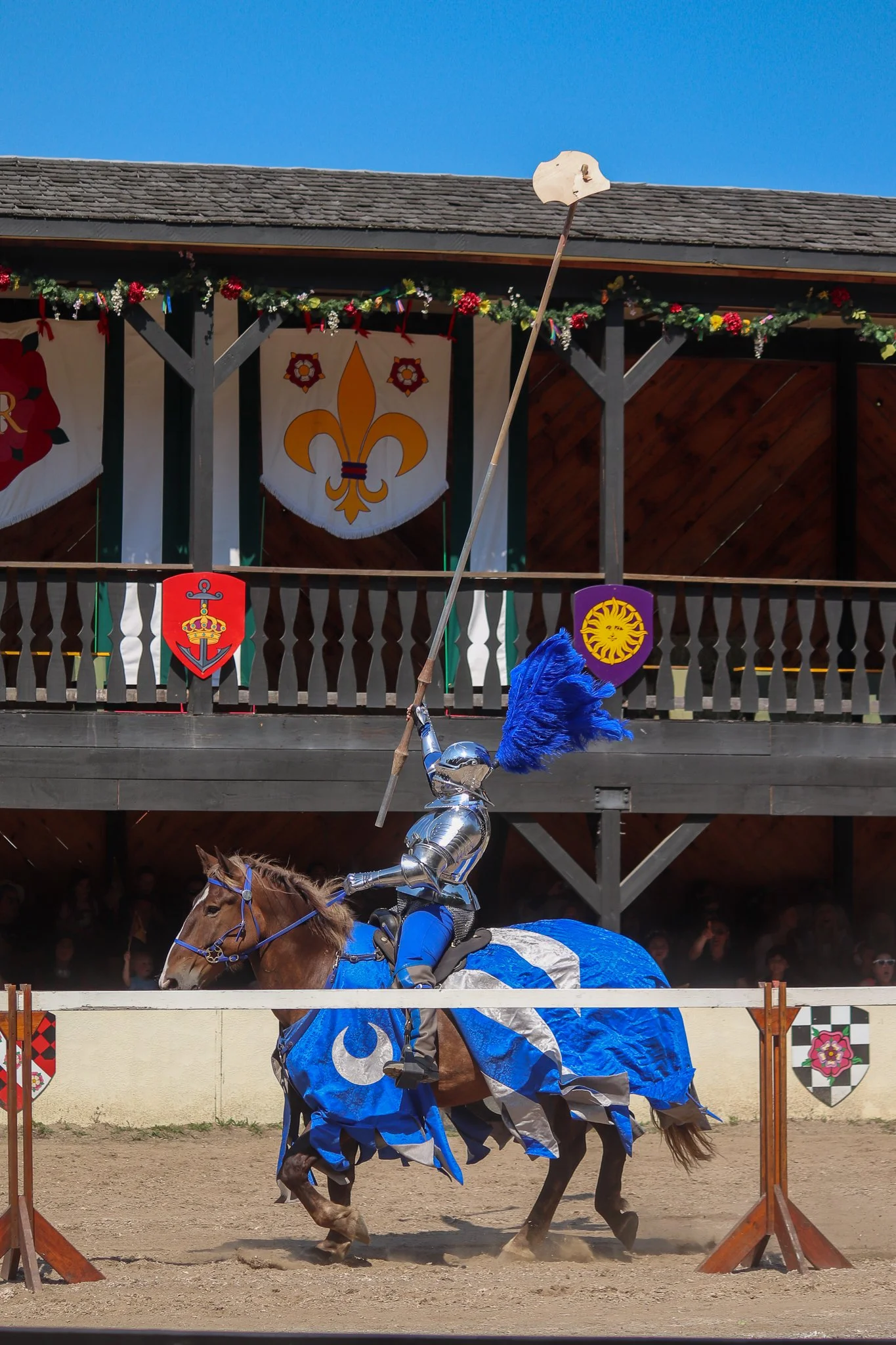 A jousting knight in armor on a brown horse dressed in a matching blue and silver caparison, holding a lance with a feathered blue plume, in a jousting arena with banners and spectators.