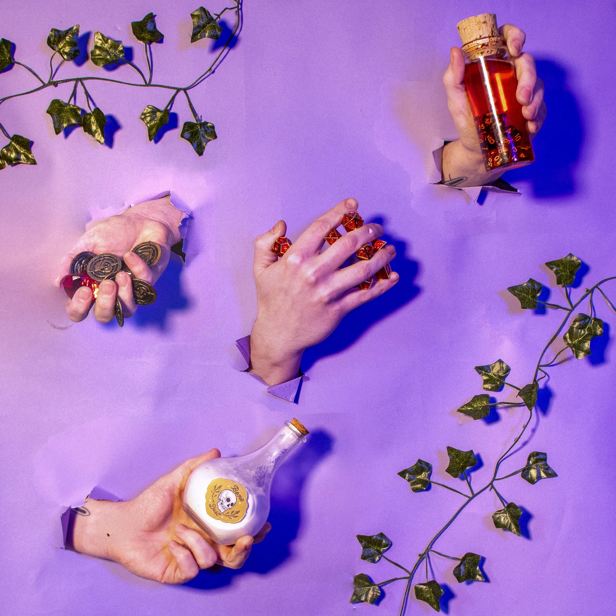 A hand holds a small glass bottle filled with a red liquid, sealed with a cork, on a purple surface. Another hand holds a small jar with a skull and floral design label. Green ivy leaves are scattered around on the purple surface.