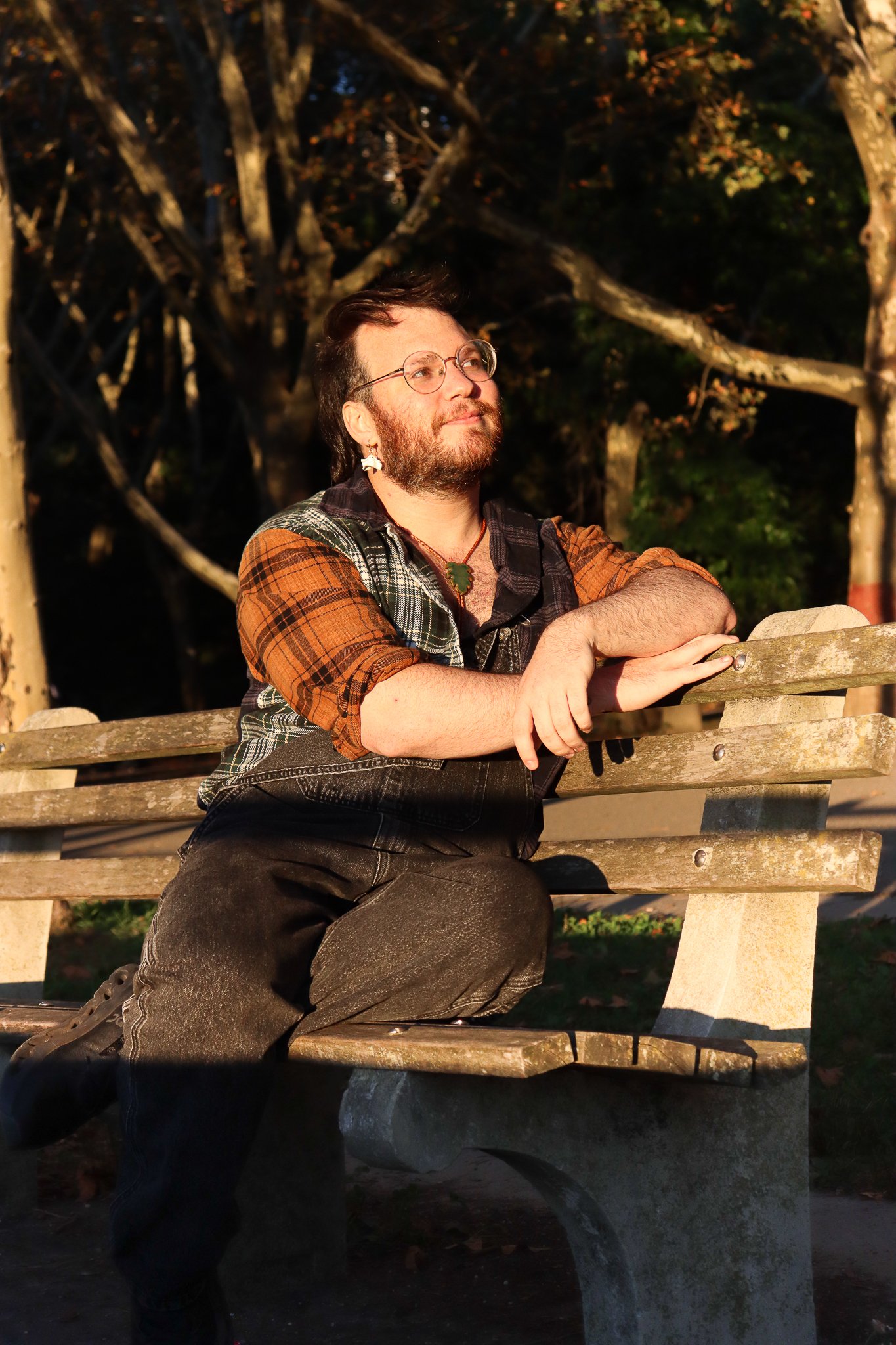 A man with glasses and a beard sitting on a wooden park bench during sunset, wearing a plaid shirt with orange and green details, surrounded by trees with leaves in autumn colors.