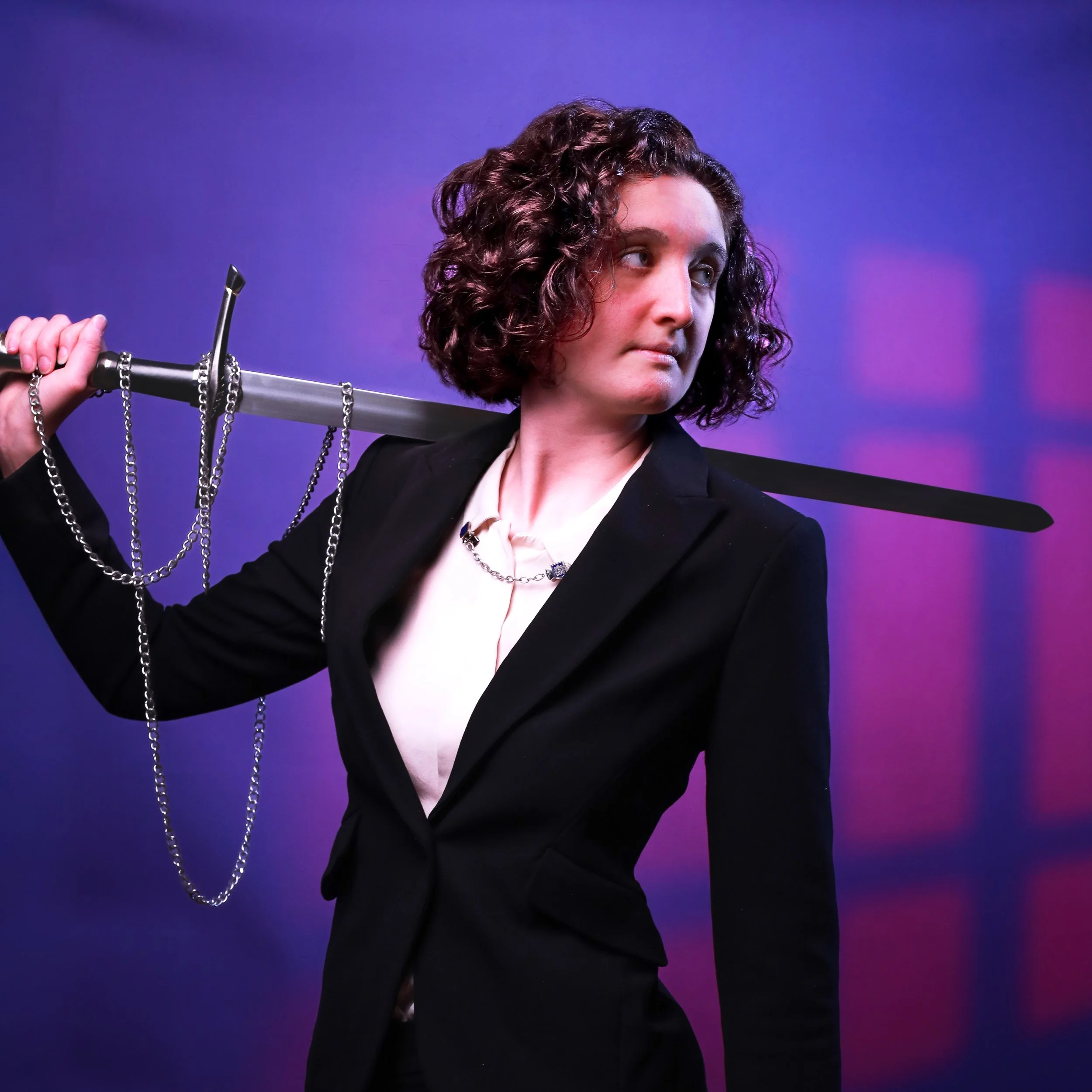 A person with curly hair wearing a black blazer and white shirt, holding a chain with a sword, standing against a purple background. Image displays high fantasy and very whimsical themes.