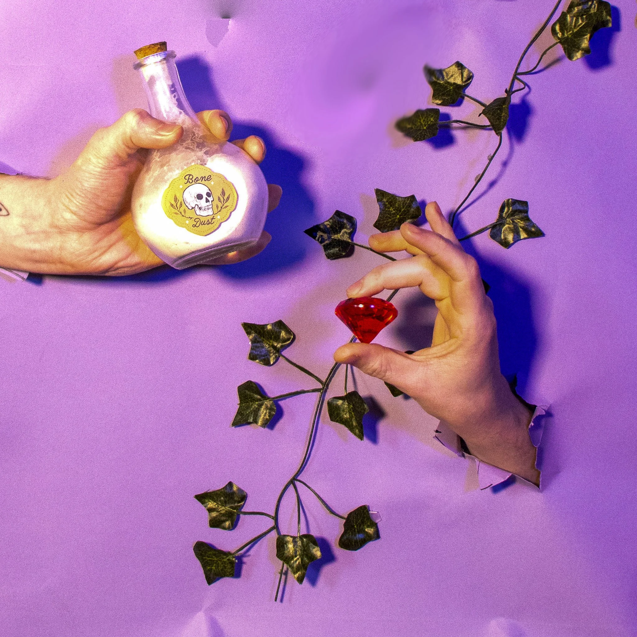 A hand holding a decorative bottle labeled 'Bone Dust' with a skull illustration, and another hand holding a leafy vine with metallic-looking leaves against a purple background.