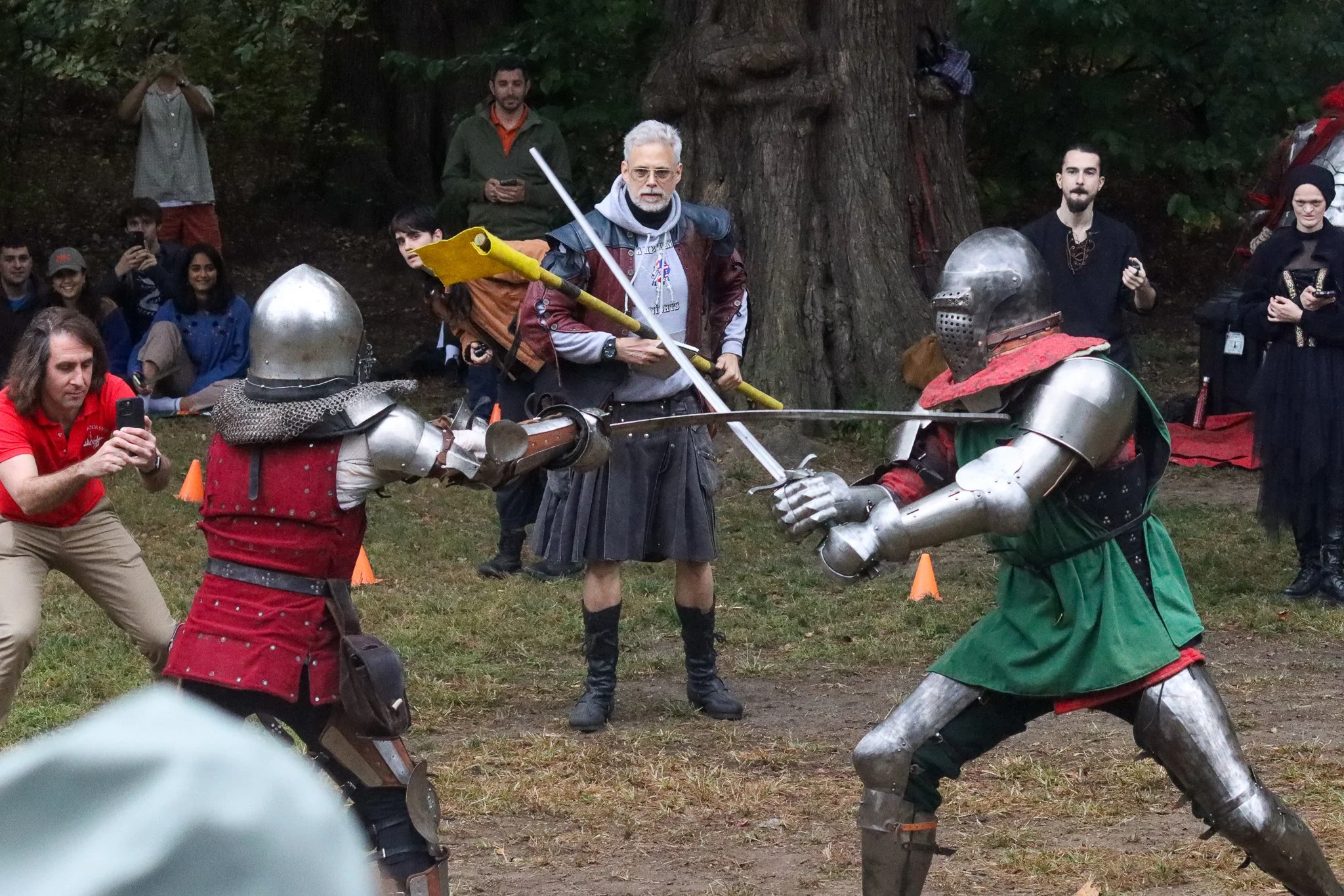 People participating in a live-action medieval combat reenactment outdoors. A man in armor and a woman with gray hair, glasses, and medieval costume are engaged in a mock fight. Spectators are watching, some taking photos or recording with their phon