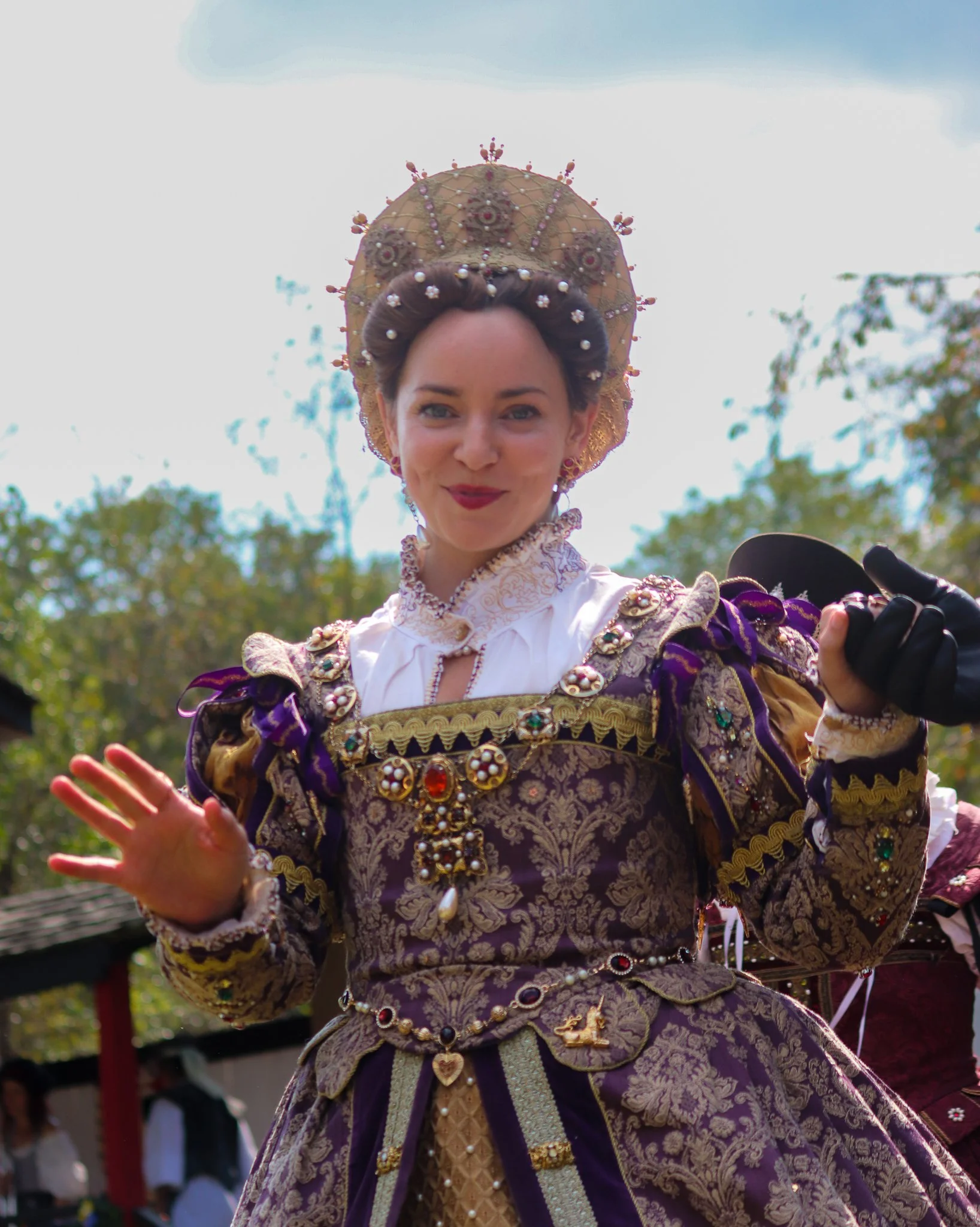 A woman dressed in a historical costume with a decorated headdress, standing outdoors with trees and a fence in the background.
