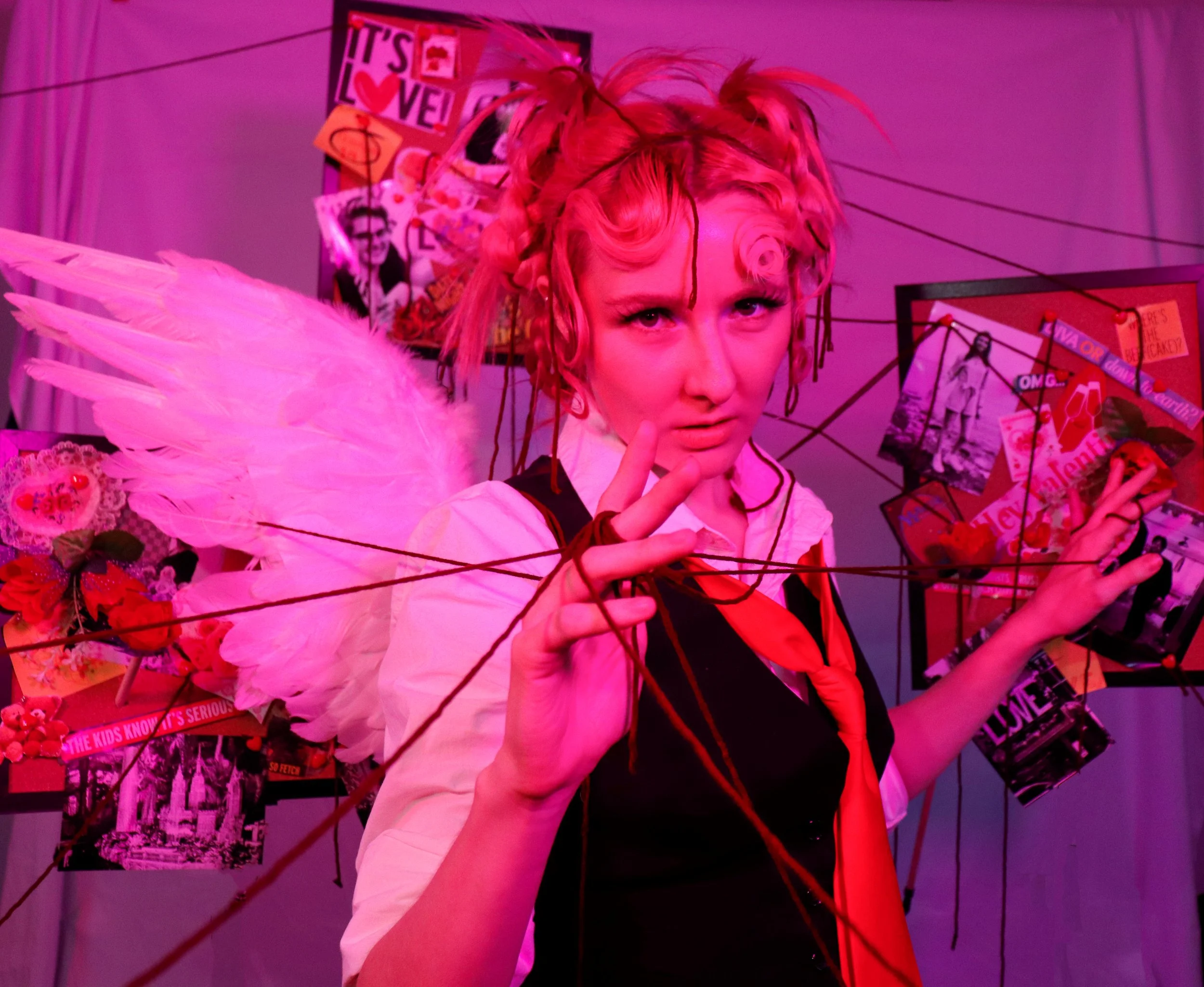 A person with short curly hair and light skin posing with their finger near their lips, wearing pink angel wings, in a room with pink lighting, and a bulletin board with various pictures, magazine clippings, and signs in the background.