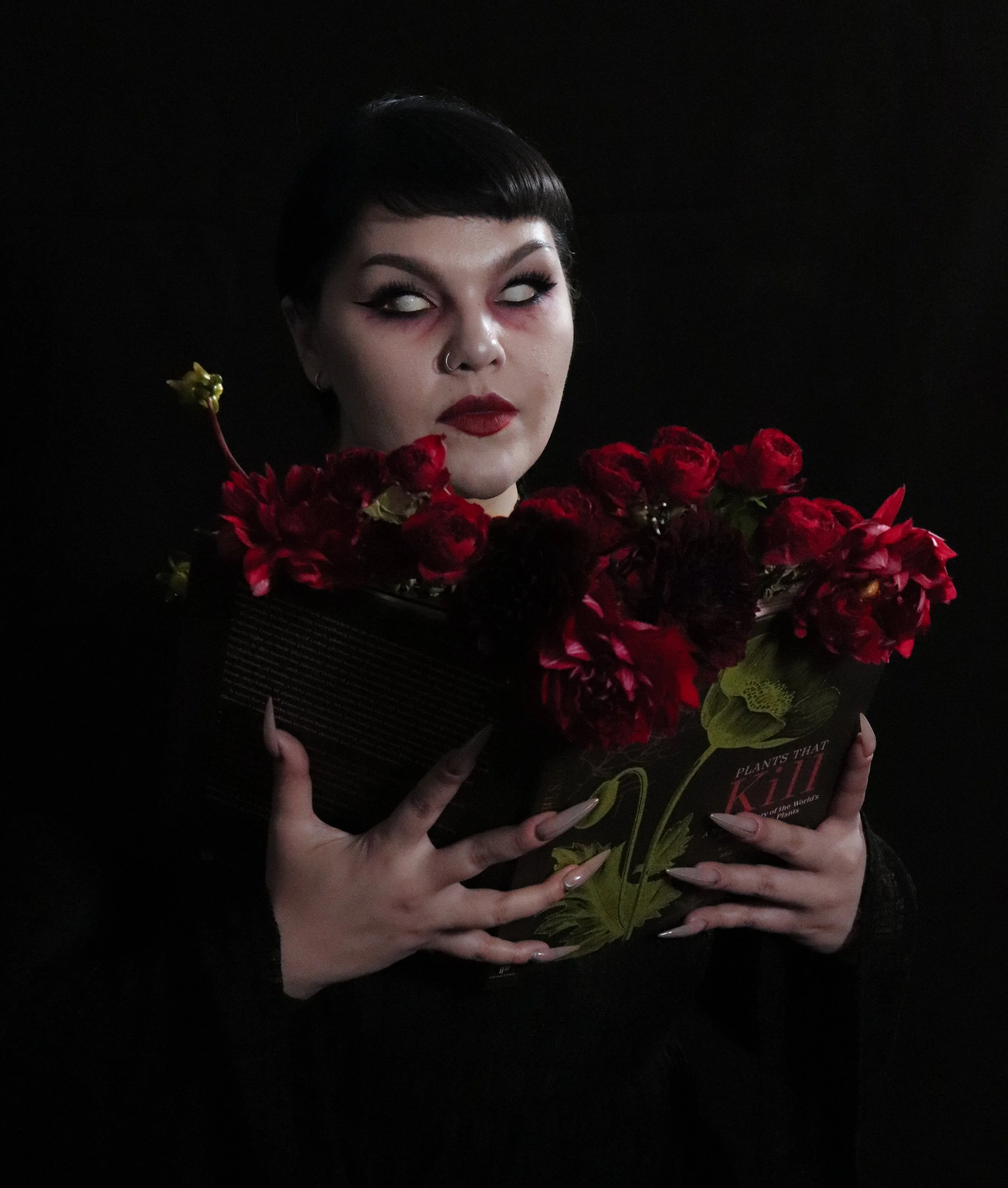 Person with short dark hair and makeup, holding a book titled 'Plants That Kill' with a bouquet of red flowers.