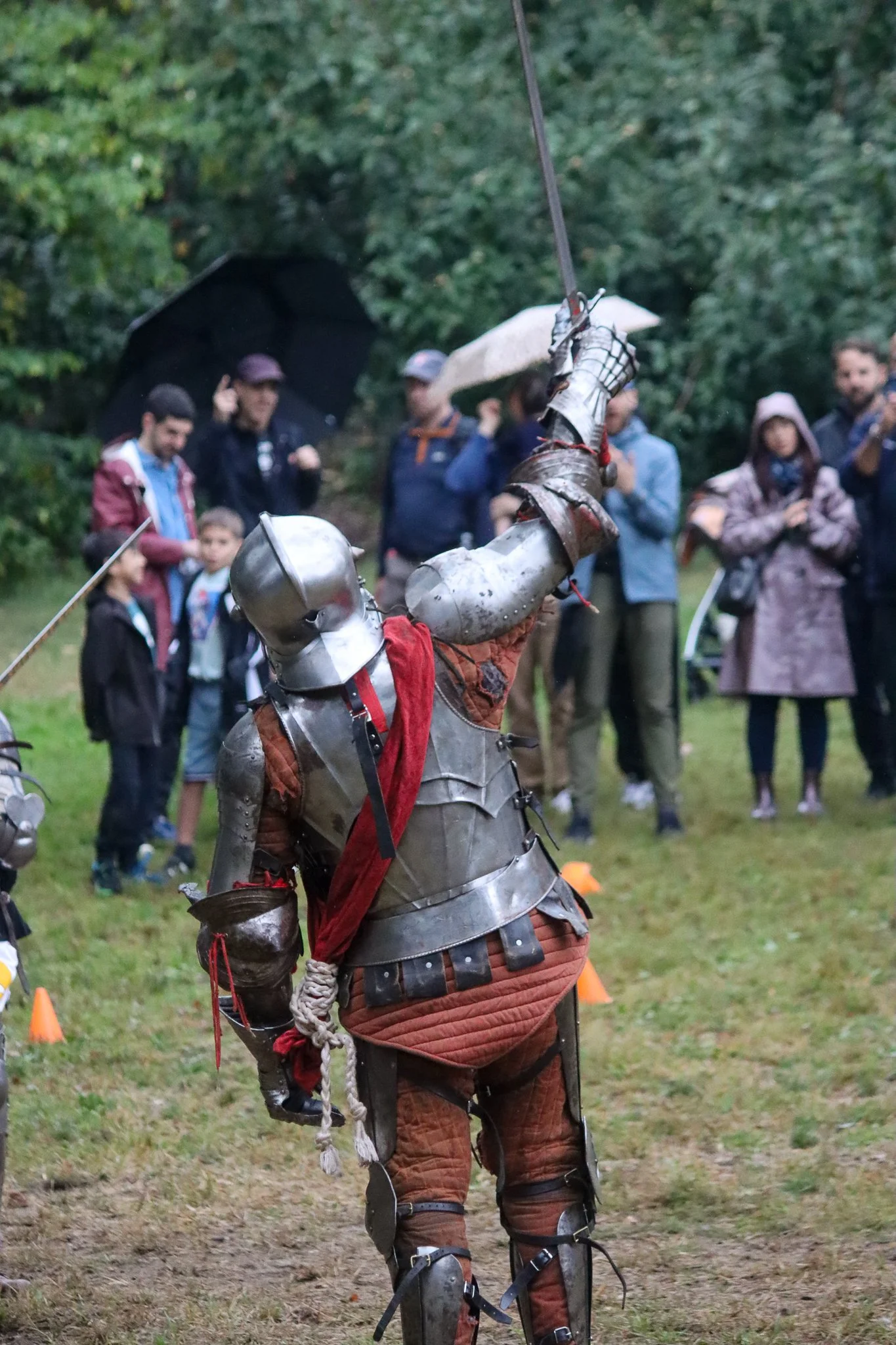 A person dressed in medieval knight armor holding a rapier in a fencing stance during an outdoor event with onlookers in the background.