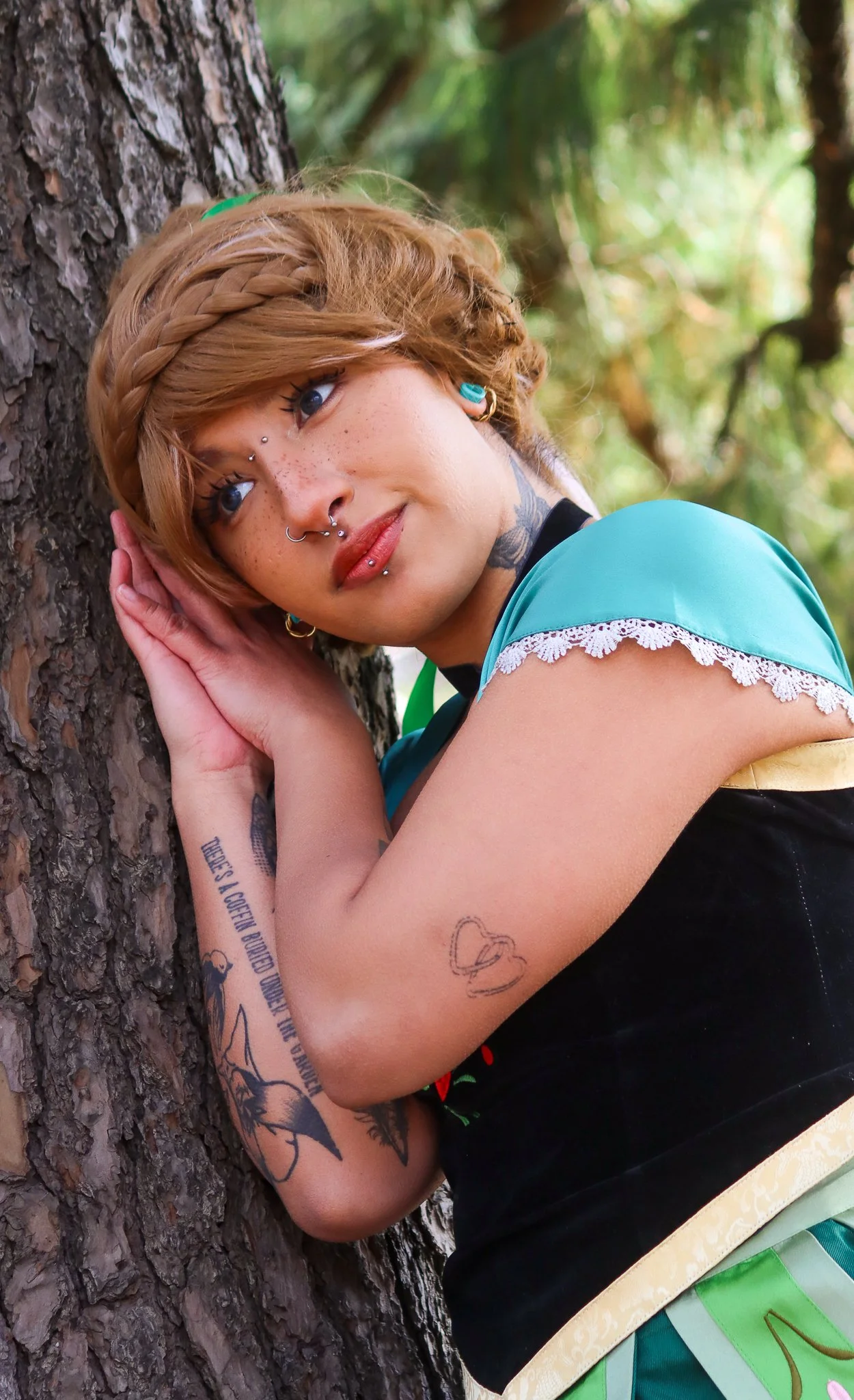 A woman with reddish-blonde hair and multiple facial piercings leaning against a tree, resting her temple on her hand in an outdoor forest setting.
