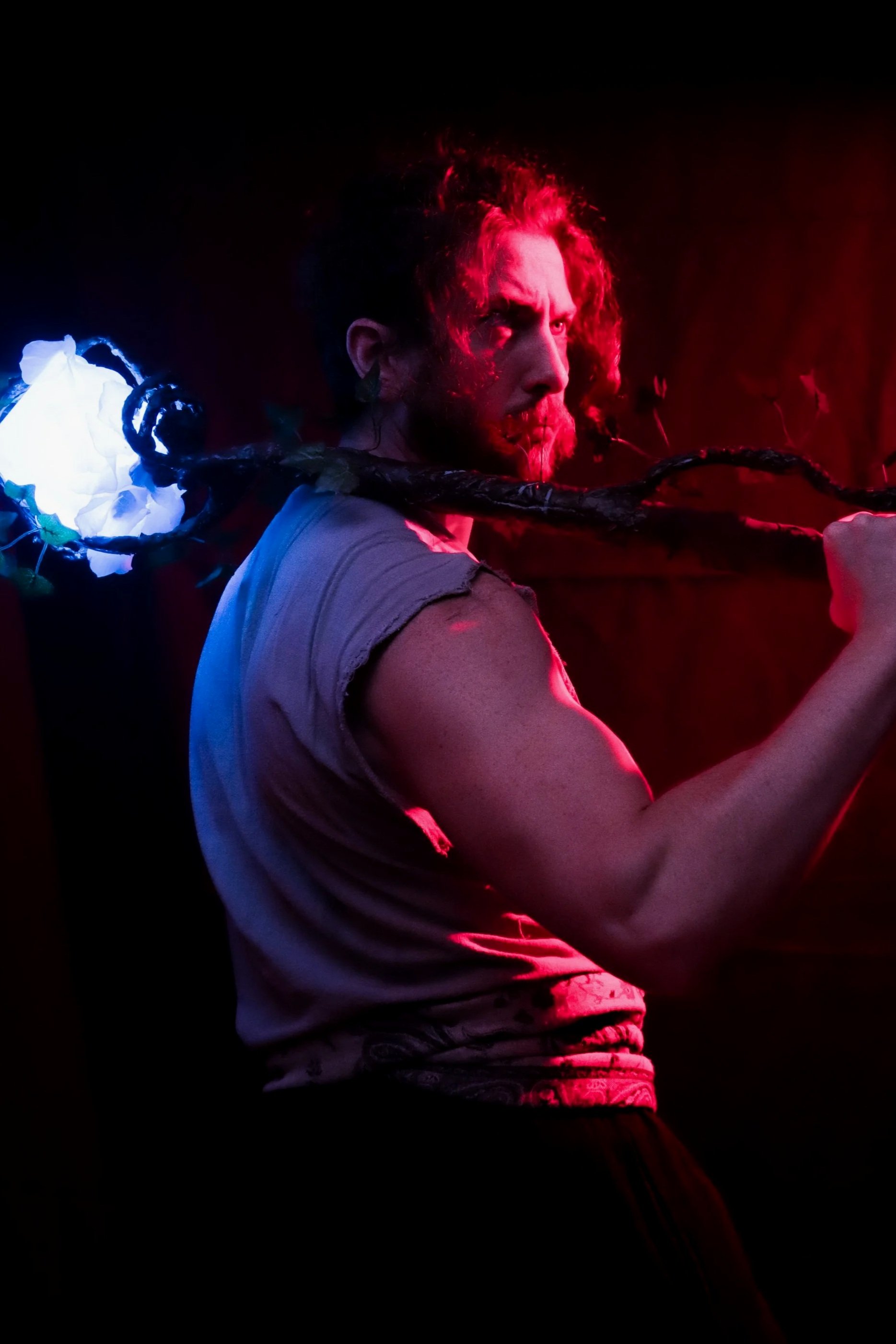 A man with curly red hair and a beard in a dark setting, with red and blue lighting highlights on his face and clothing, holding a long object over his shoulder.