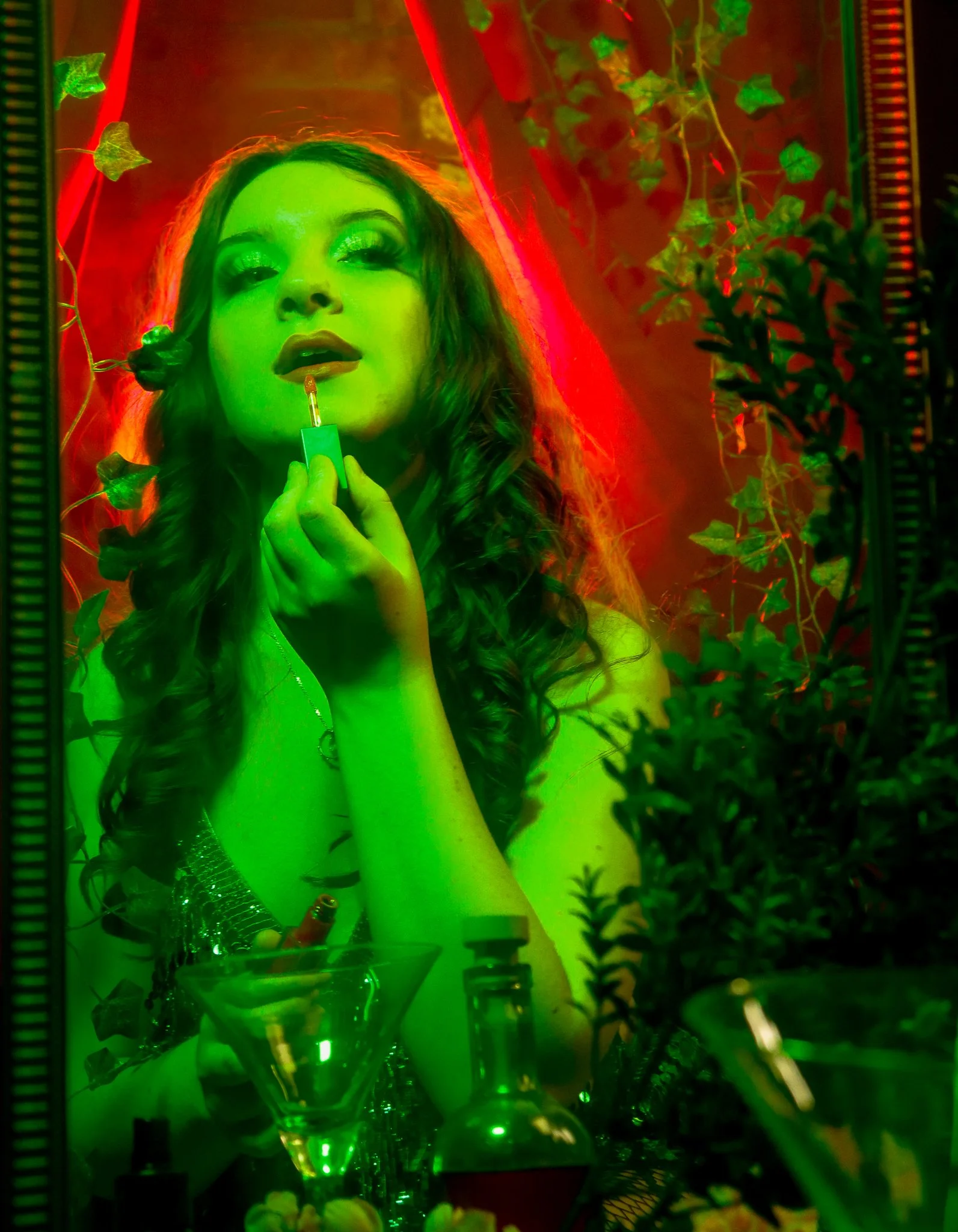 A woman with long wavy hair, glittery makeup, and lipstick applying lipstick in front of a mirror illuminated with green light. The background has red lighting and some green leafy vines.