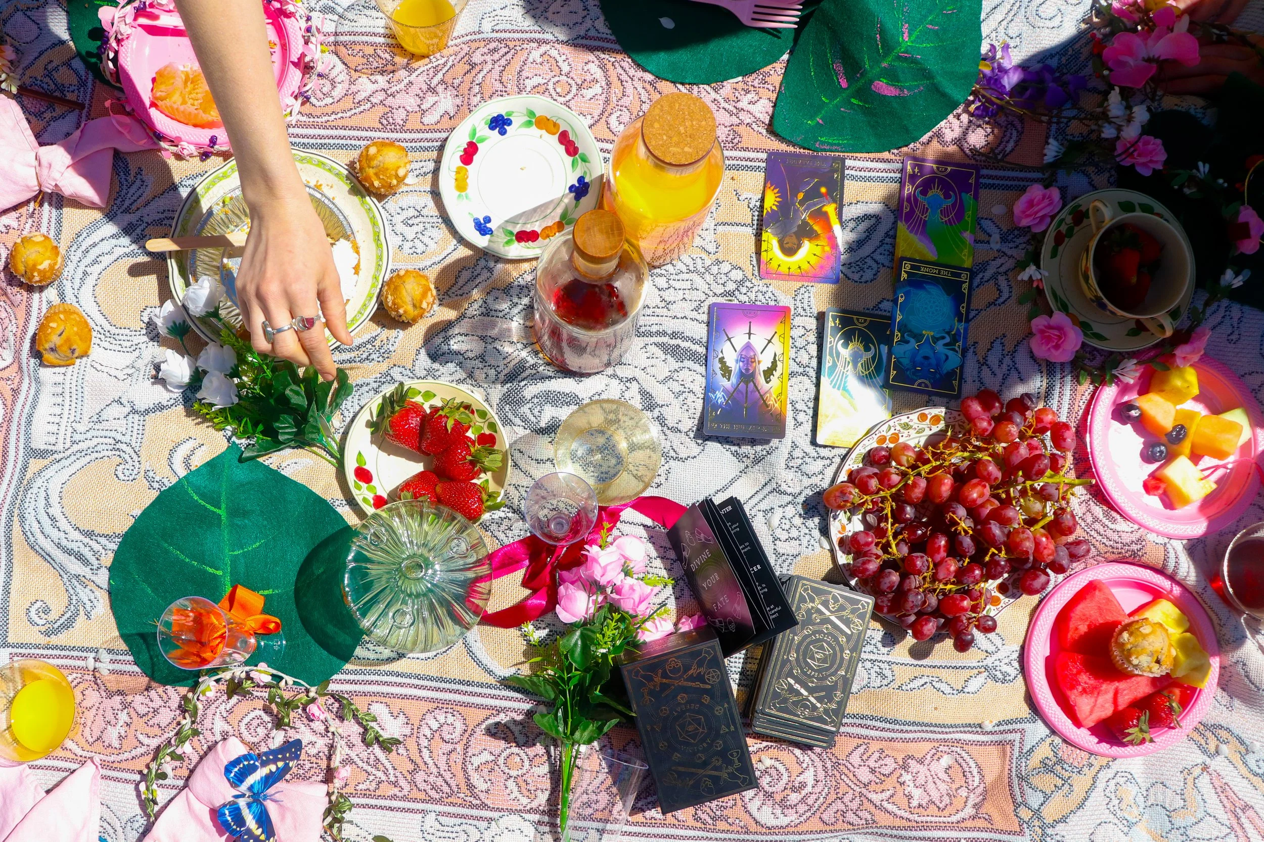An assortment of items on a decorated outdoor table, including tarot or oracle cards, colorful flowers, fruit, drinks, and small dishes, set for a daytime gathering or celebration.