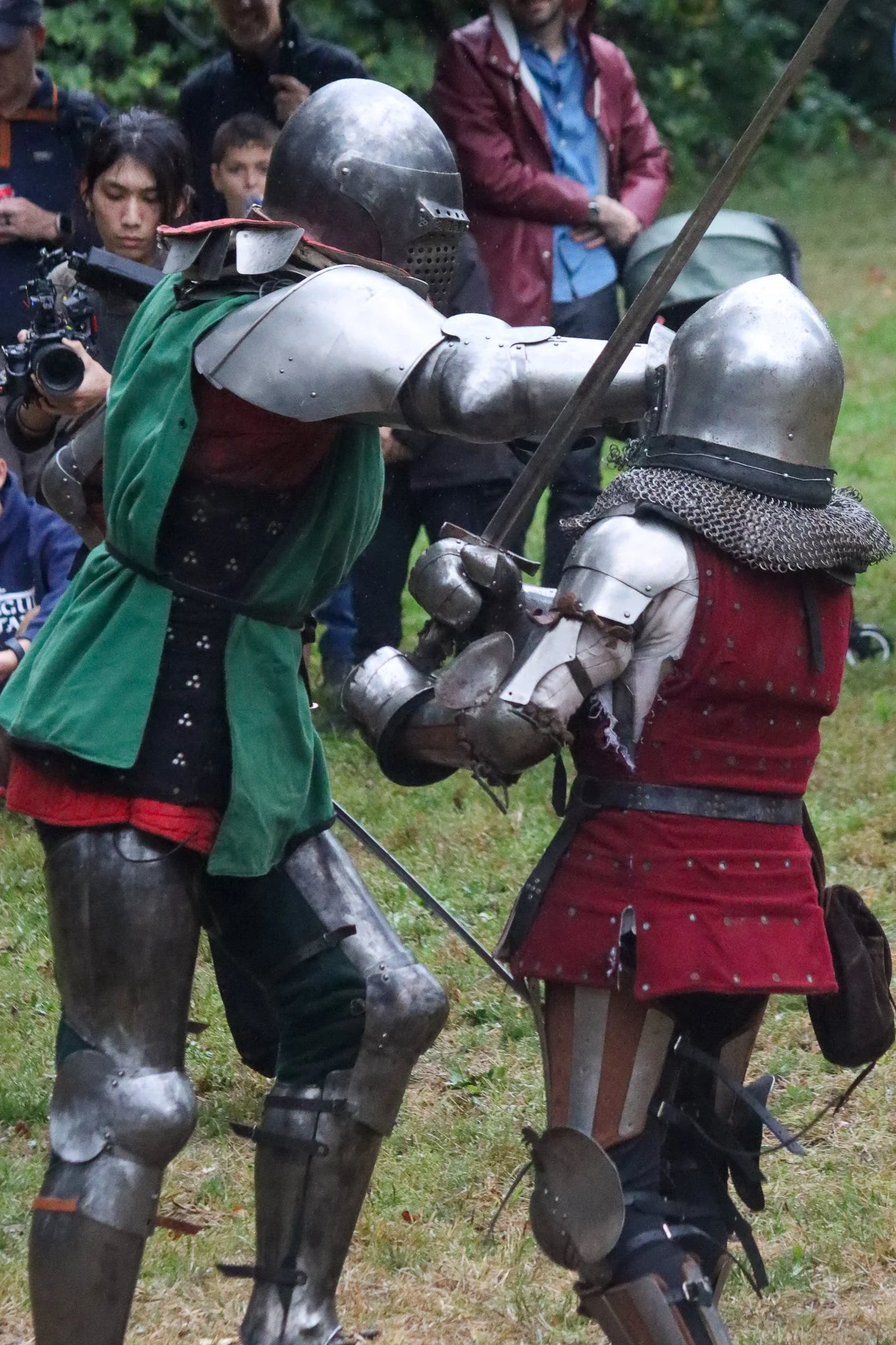 Two people dressed in medieval armor are engaged in a sword fight, surrounded by onlookers in casual clothing in an outdoor setting.