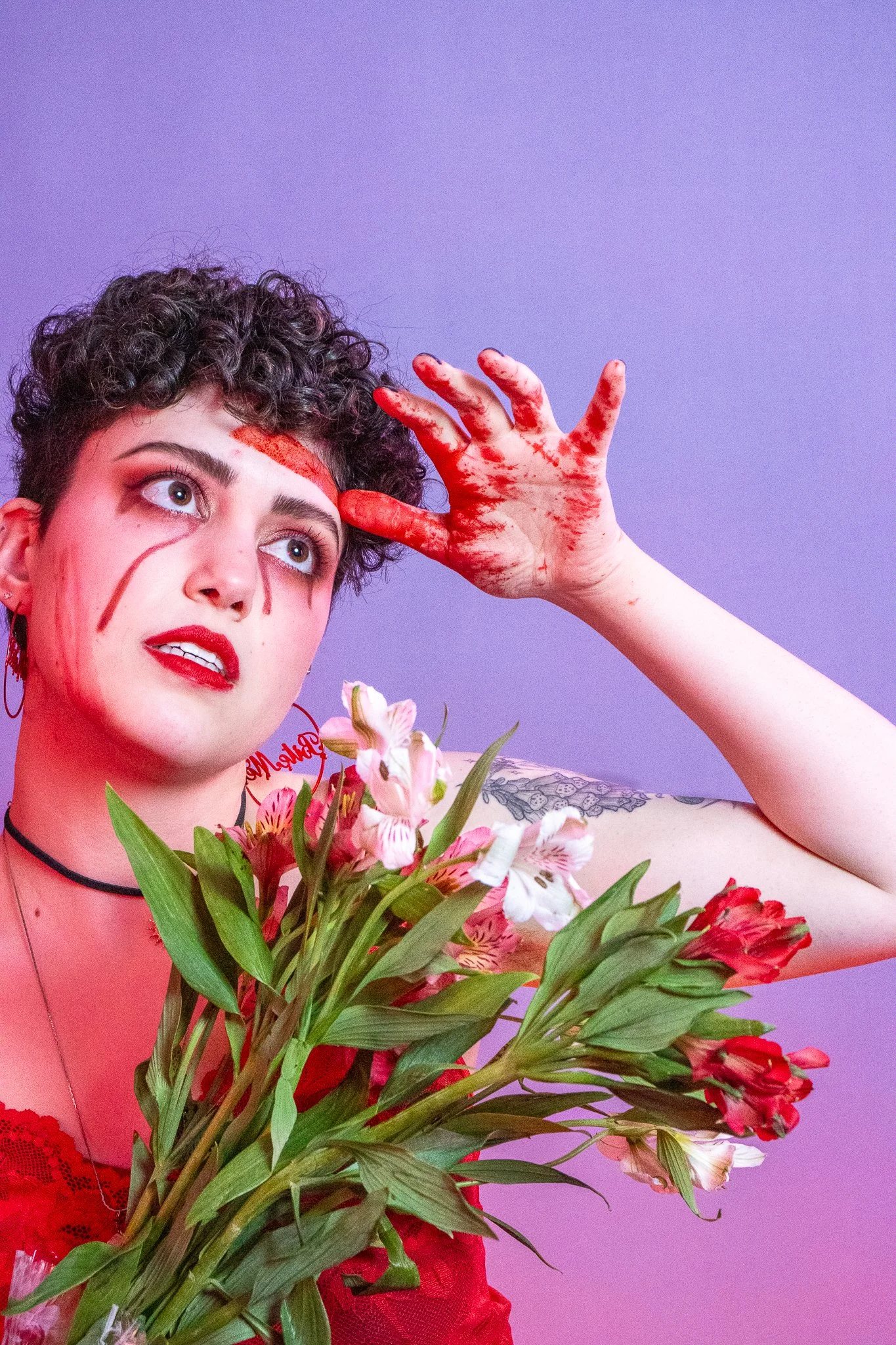 Person with short curly hair holding colorful flower bouquet, nose and face painted with red makeup, wearing red lace clothing, standing against purple background.
