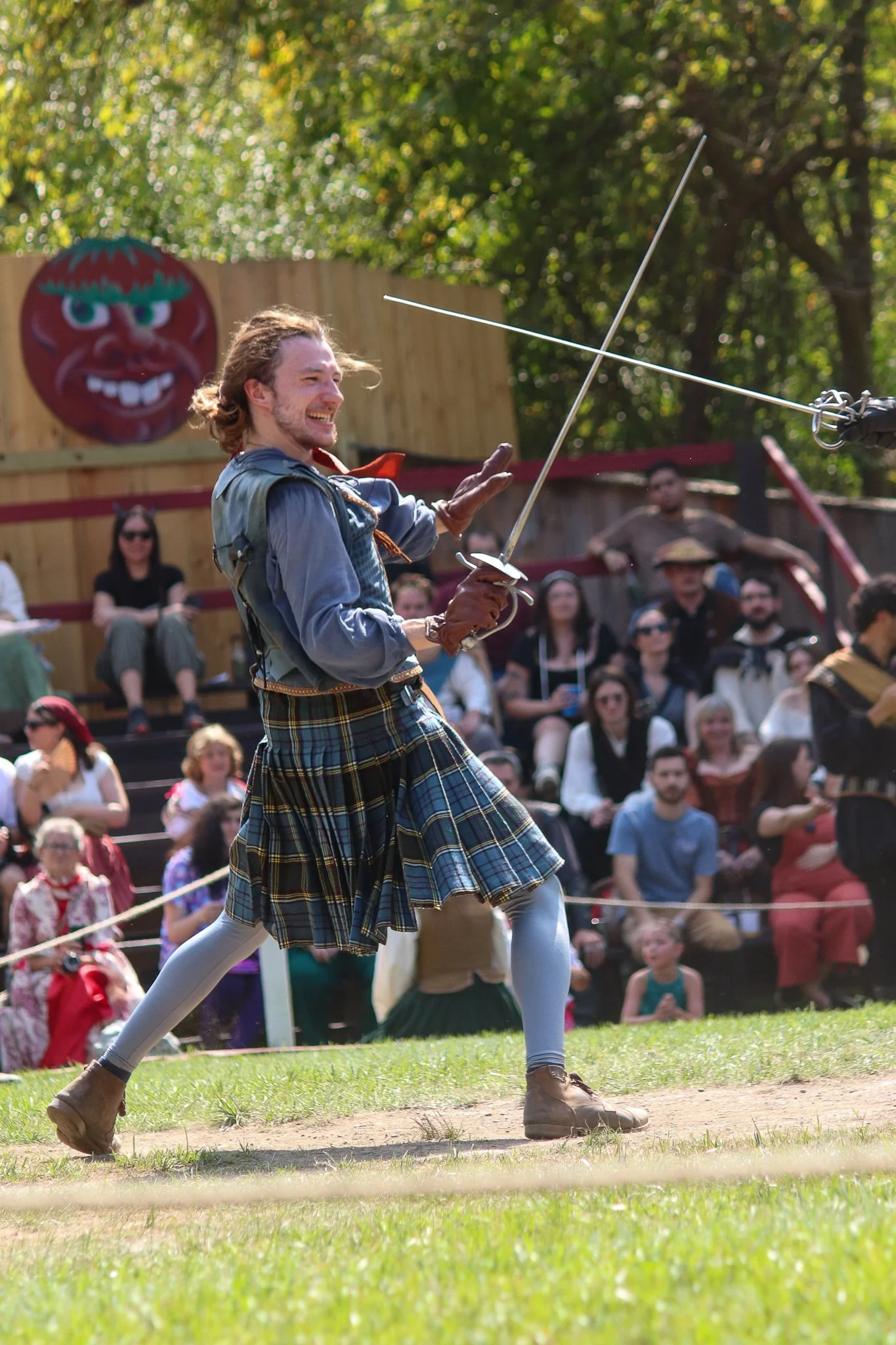 A man dressed in Scottish attire, including a kilt, participating in a sword dance outdoors with an audience watching.
