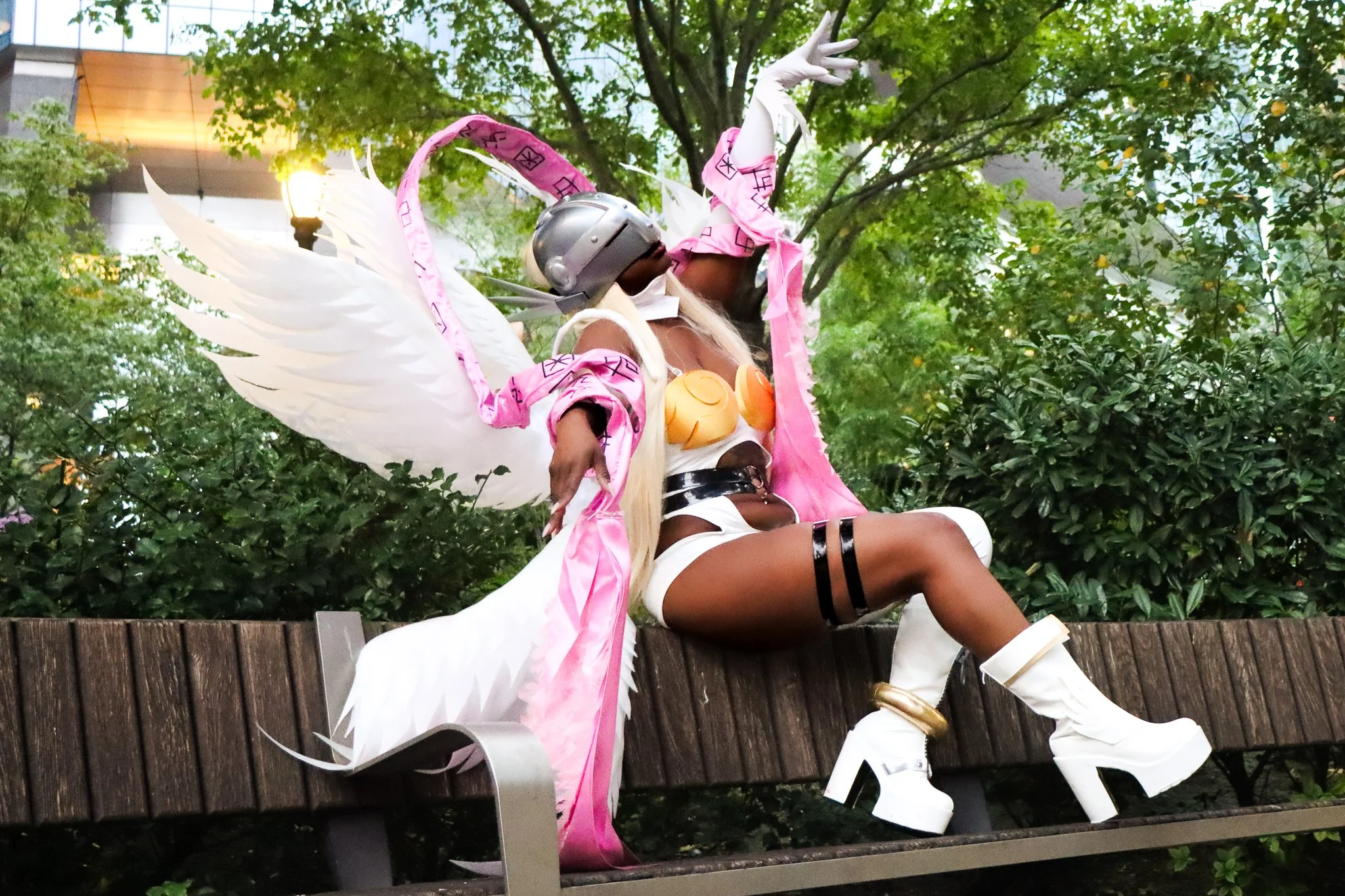 Person dressed in a fantasy costume with white wings, a pink and white outfit, and a silver helmet, sitting on a park bench among green trees.