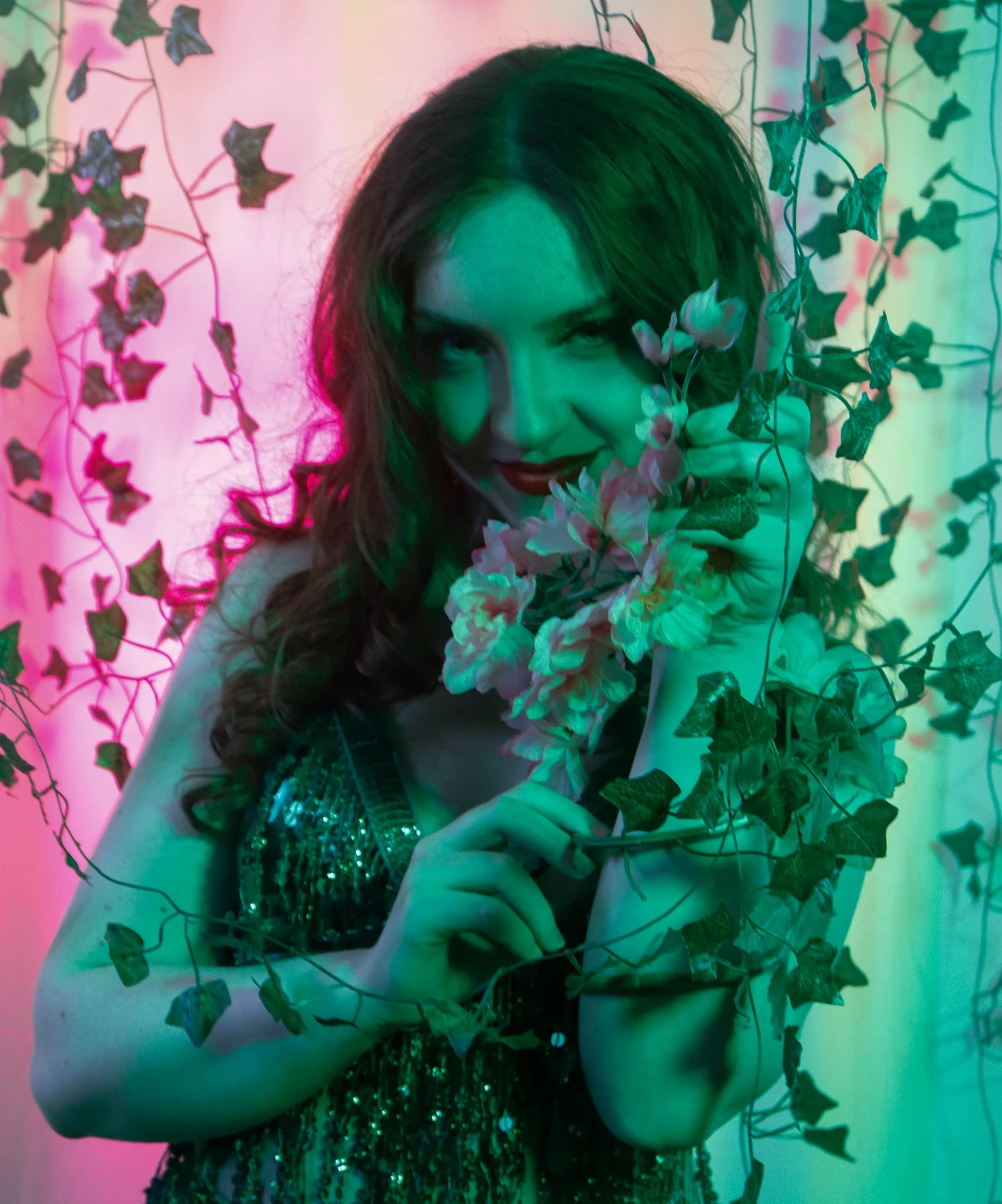 A woman with long brown hair, wearing a shiny, sequined dress, holding pink and white flowers, and surrounded by leafy vines with multicolored lighting.