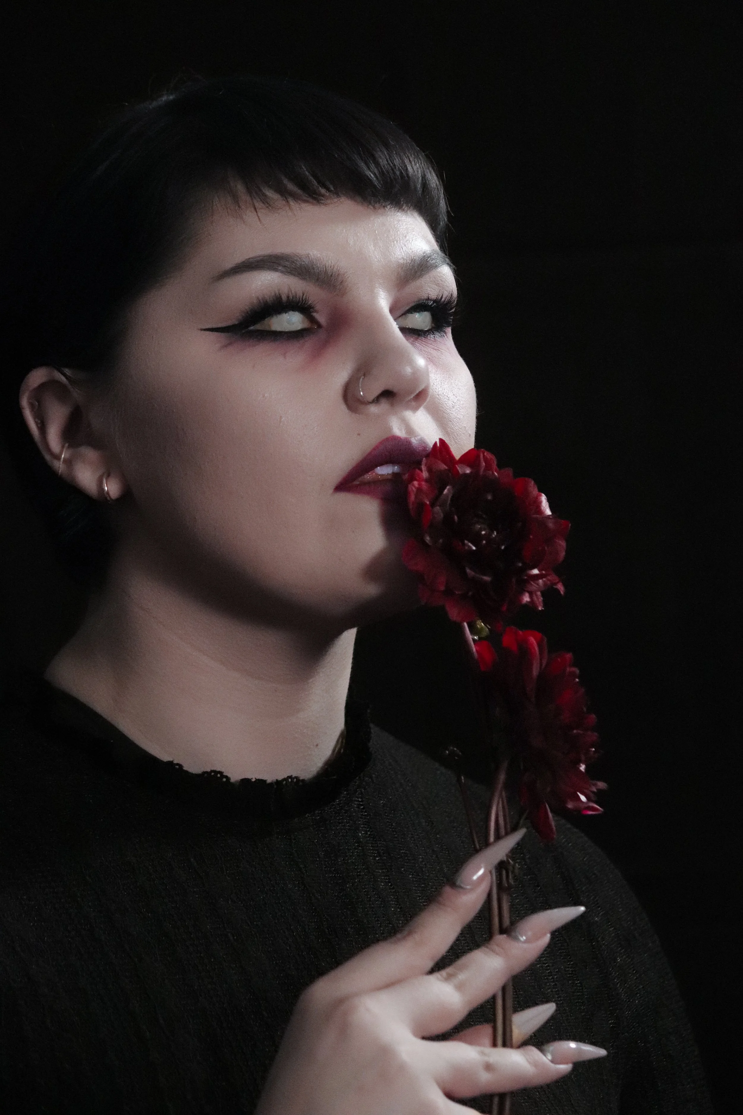 A woman with short dark hair and makeup holding a red flower close to her lips against a black background.