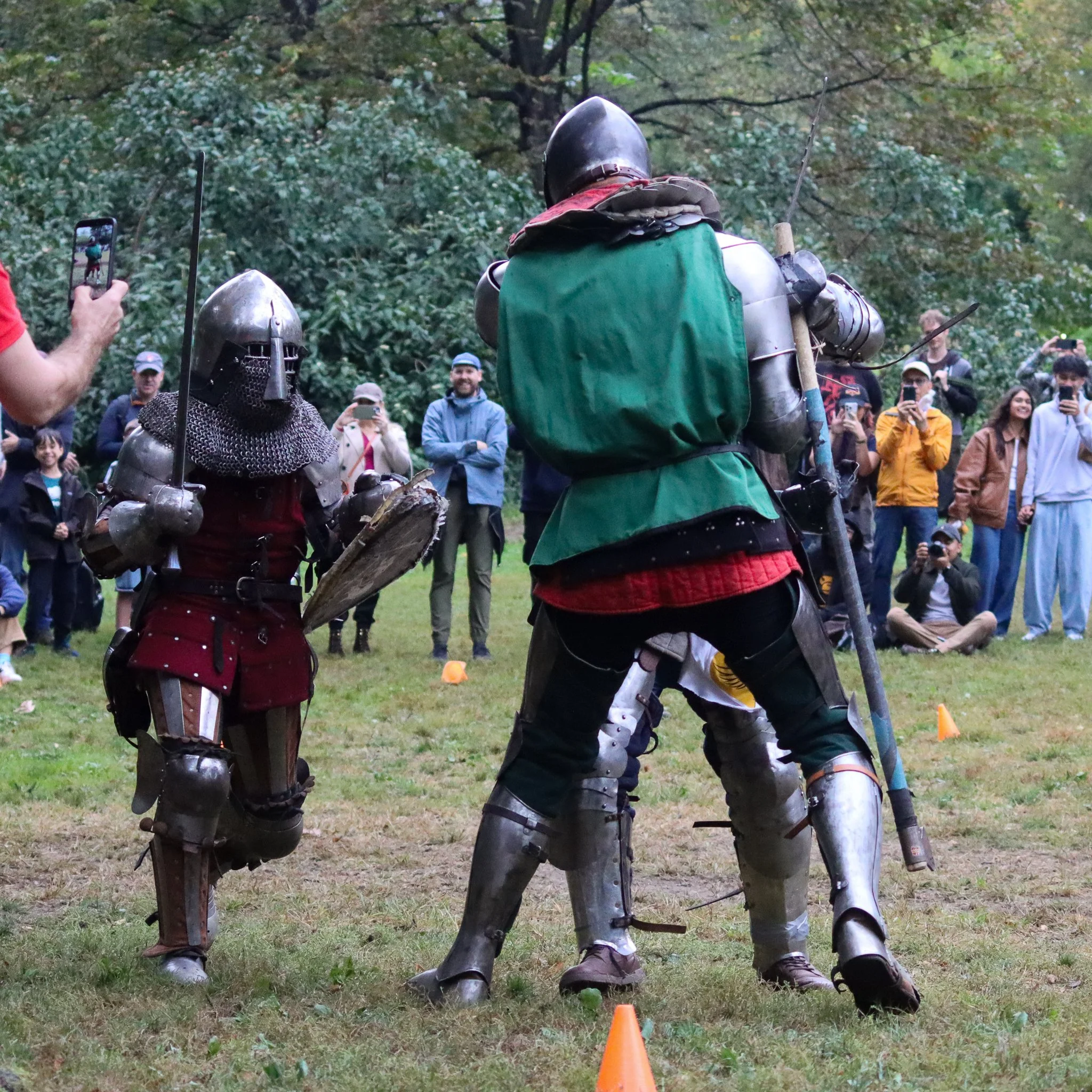 Two individuals dressed as medieval knights in armor facing each other, with onlookers observing and taking photos in the background, in an outdoor setting with trees.