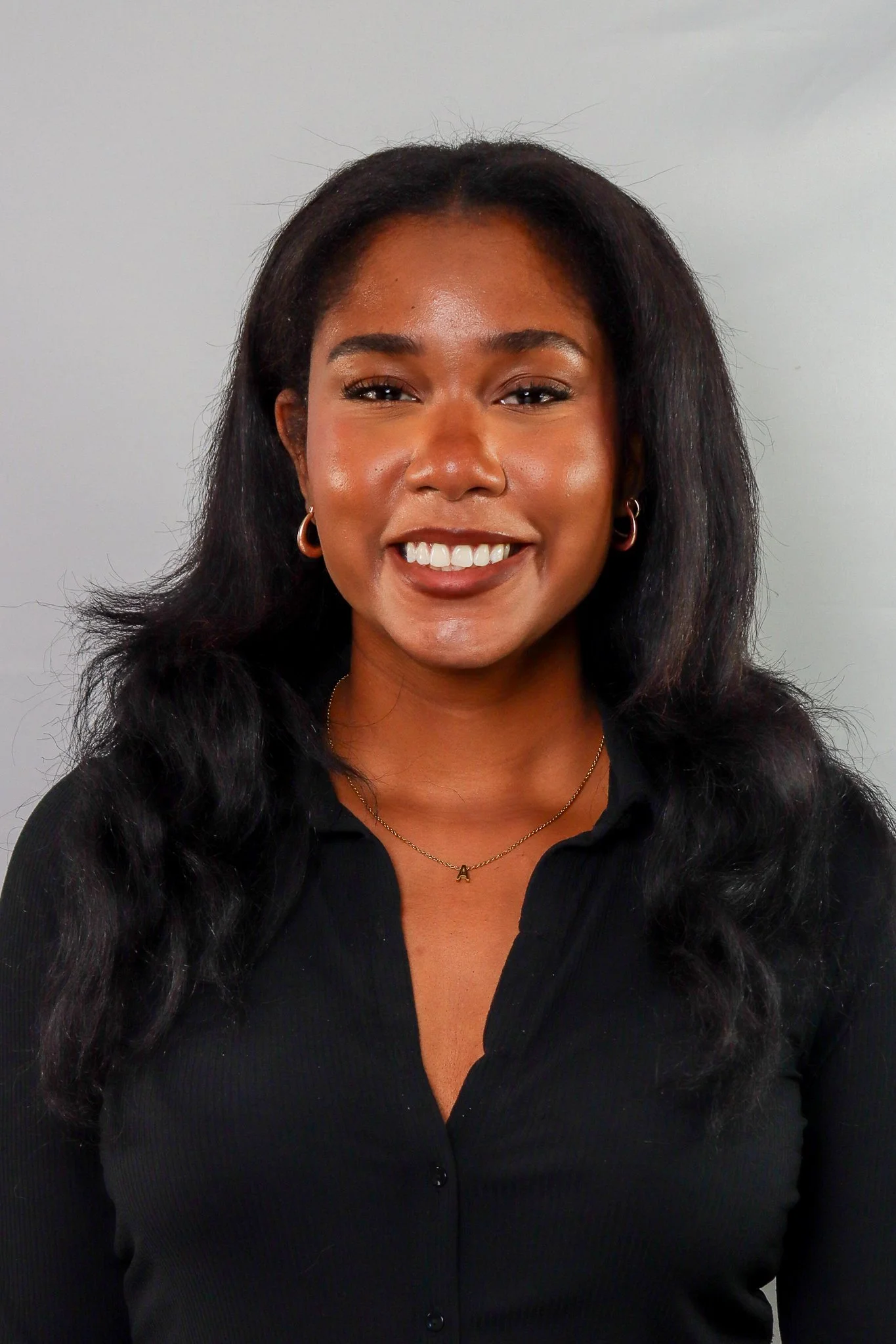 A woman with long, wavy black hair smiling, wearing hoop earrings, a gold necklace with a pendant, and a black button-up top, standing against a plain light gray background.