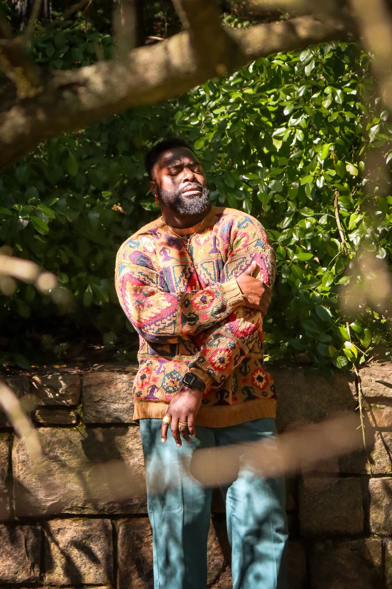 A man with dark skin, a beard, and short hair is posing outdoors among green leafy bushes and a stone wall. He is wearing a colorful patterned sweater, light blue jeans, and accessorized with rings, earrings, and a black watch, with his arms crossed 