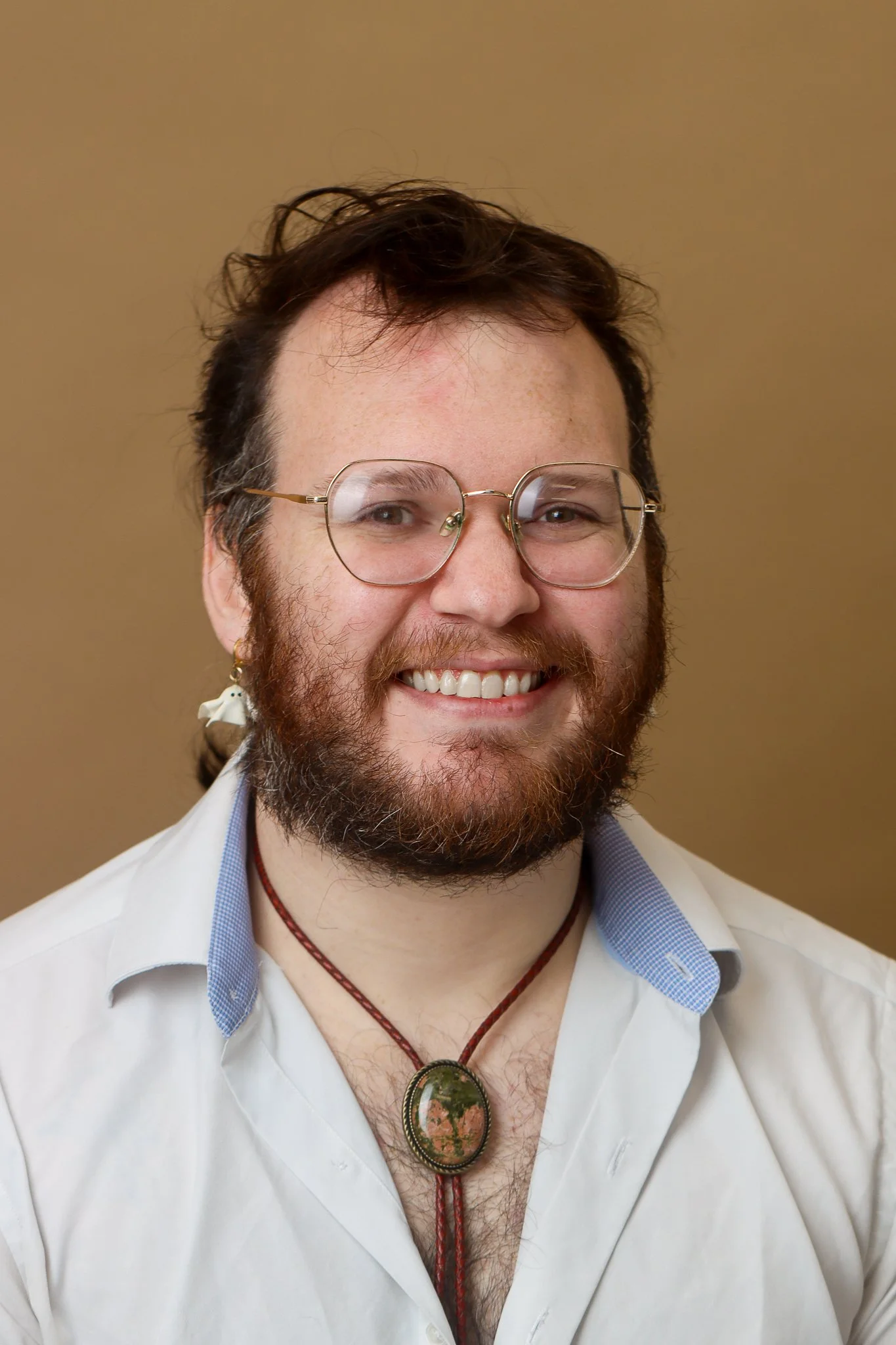 A smiling man with glasses and a beard, wearing a white shirt, a necklace with a large pendant, and an earring, against a plain background.