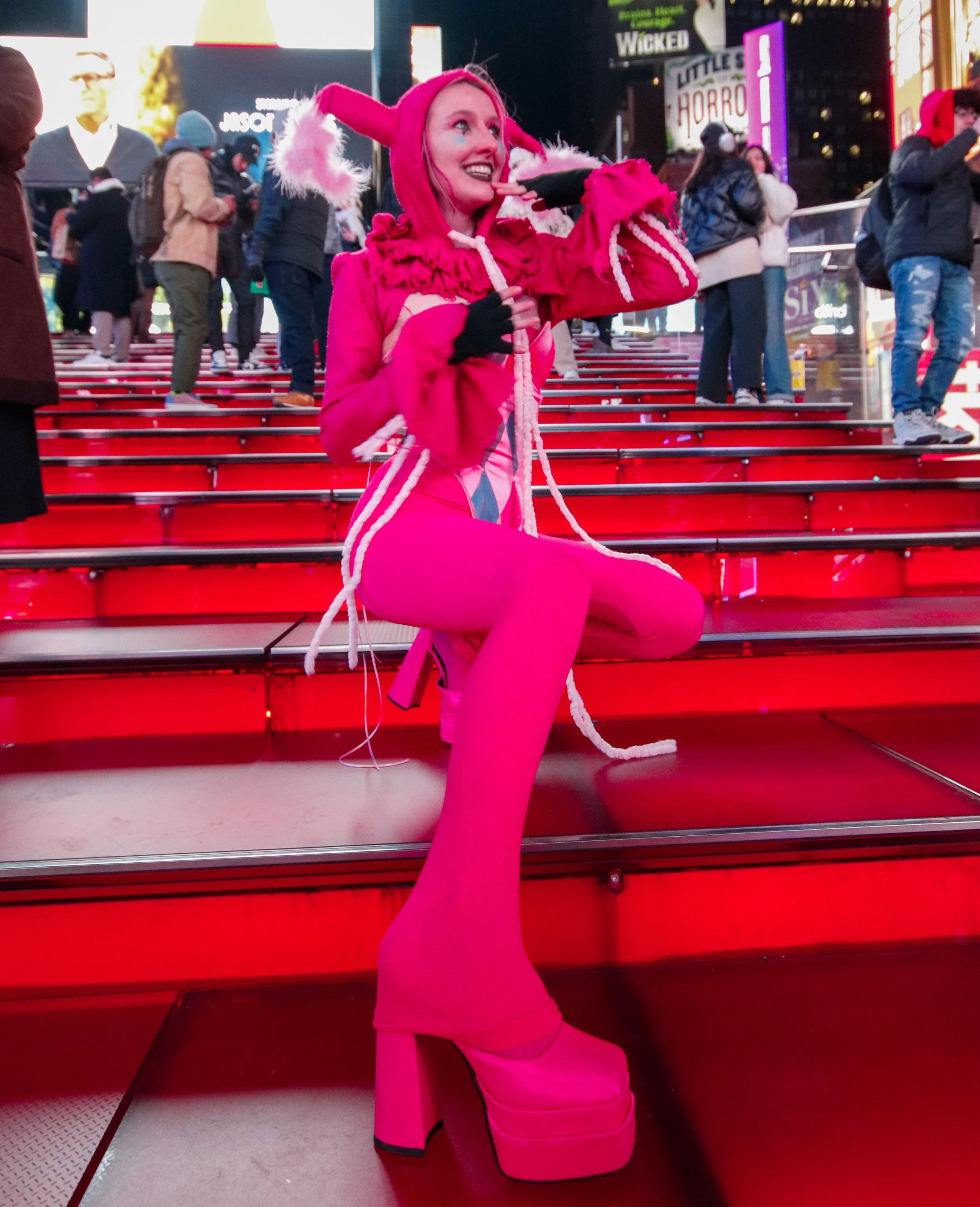 Person dressed in a bright pink costume with a hood that has bunny ears, sitting on the steps of a busy city square illuminated by neon lights.