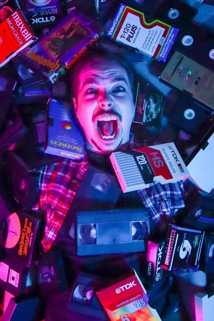 A person lying on the floor surrounded by various old technology items and VHS tapes, with a large printed face of a young man making an excited or surprised expression at the camera.