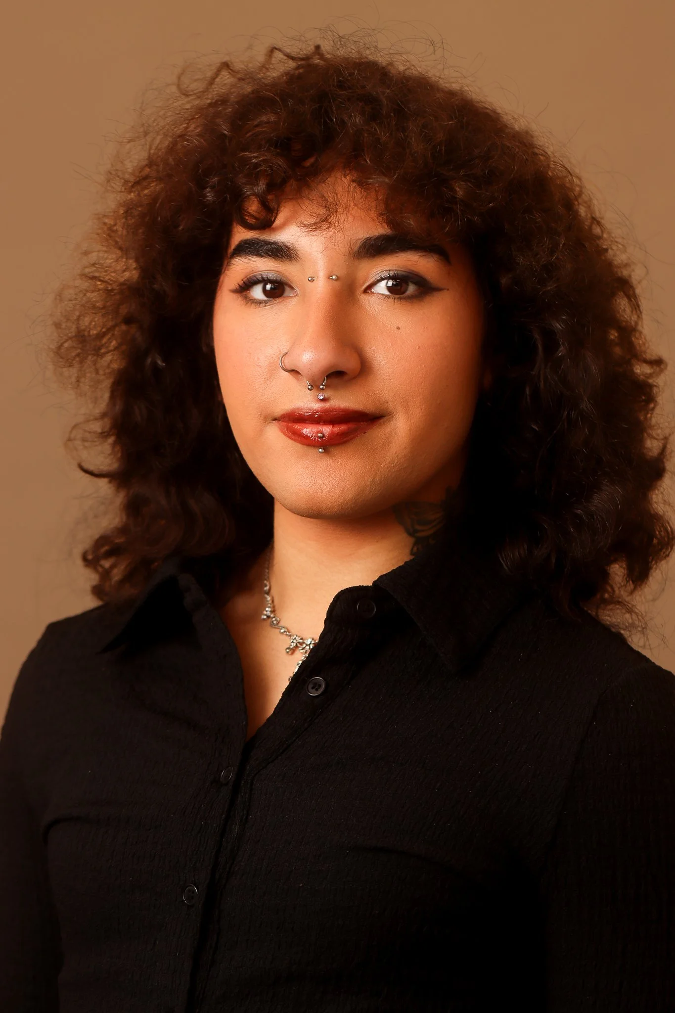 A young woman with curly hair, multiple facial piercings, and wearing a black button-up shirt, standing against a neutral background for their headshot.