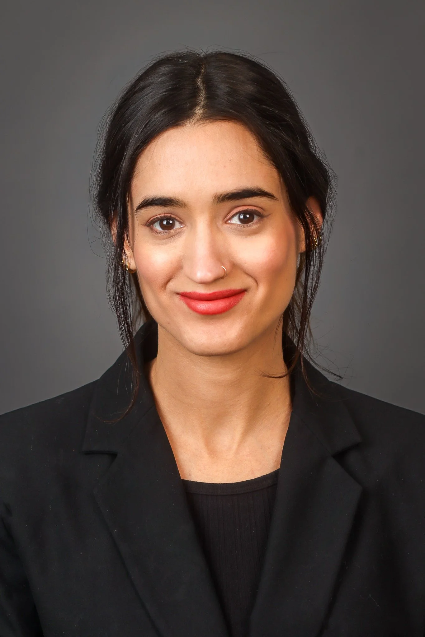 A woman with dark hair styled in loose waves, wearing a black blazer and a black top, smiling against a gray background.