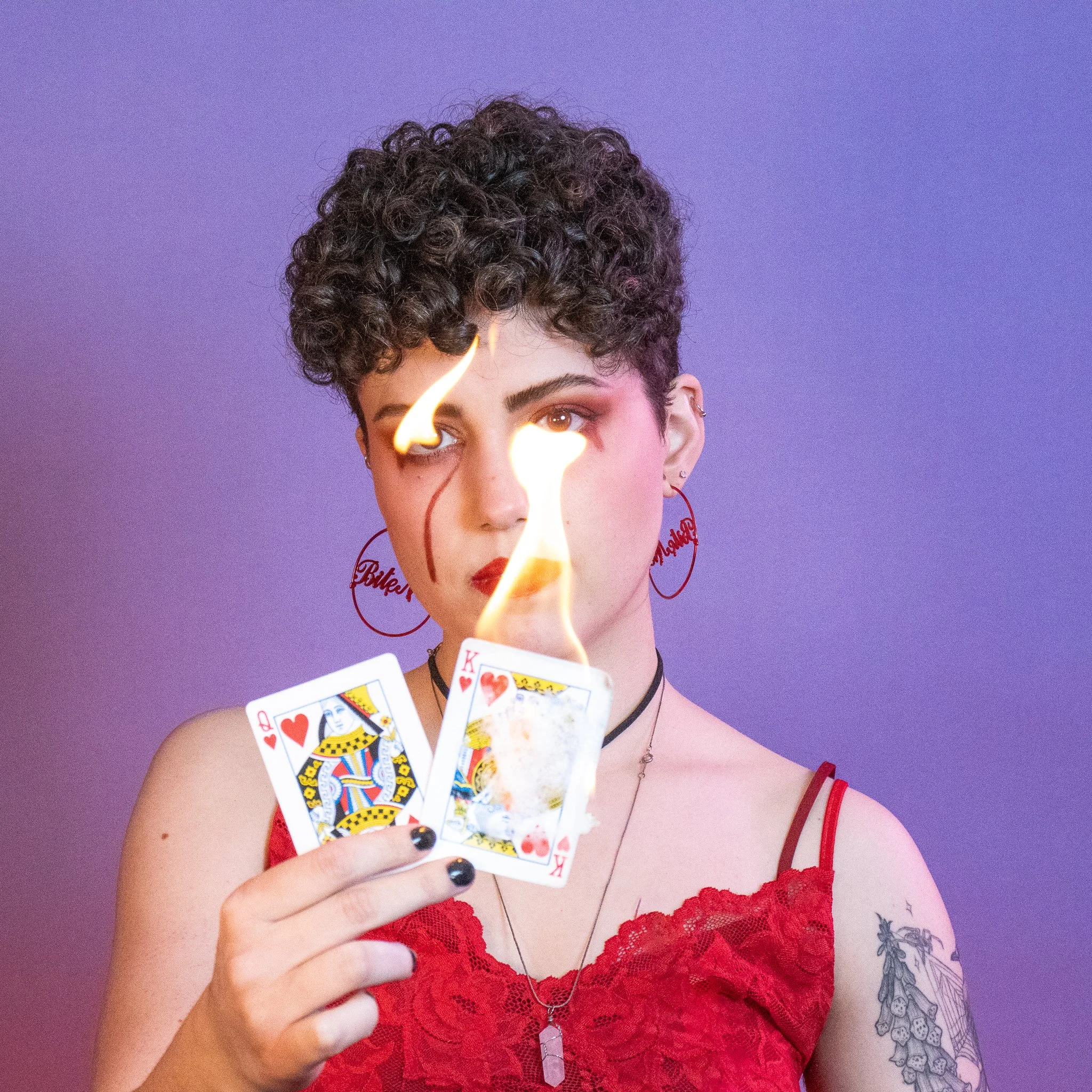 A woman with short, curly dark hair, wearing red lace lingerie, holding playing cards, with flames in front of her face, against a purple background.