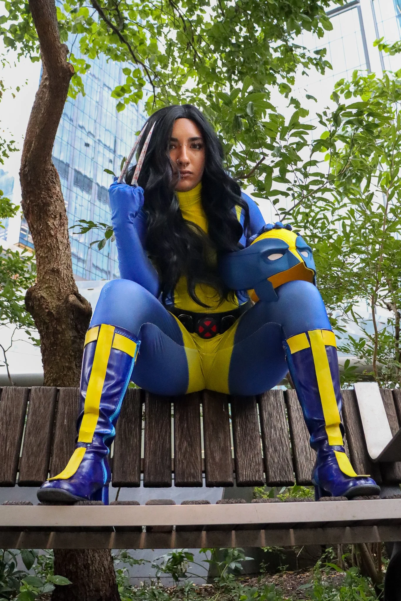 A young woman with long black hair, dressed as a superhero in blue and yellow costume, sitting on a wooden fence in an outdoor setting with trees and modern glass buildings in the background.