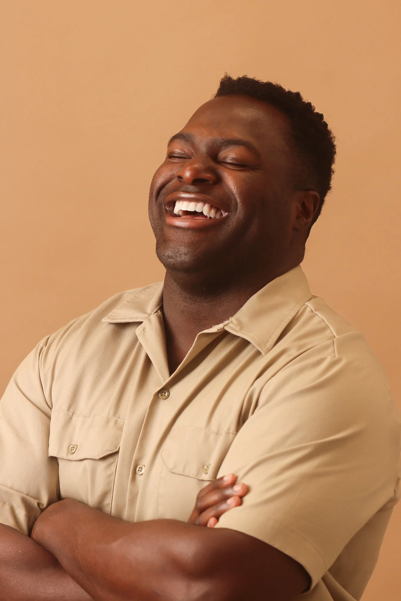 A man in a beige button-up shirt smiling with eyes closed and arms crossed, standing against a plain beige background.