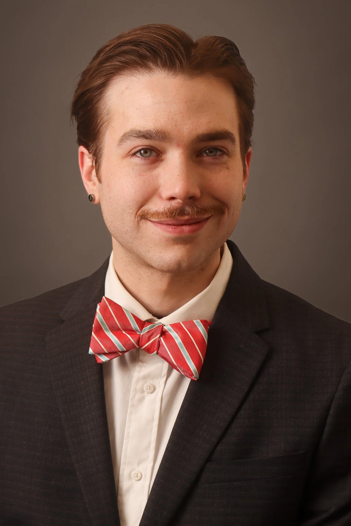 A young man with light skin, slicked-back brown hair, and blue eyes, wearing a formal black suit, a white dress shirt, a colorful striped bow tie, and a stud earring, smiling confidently against a plain gray background.