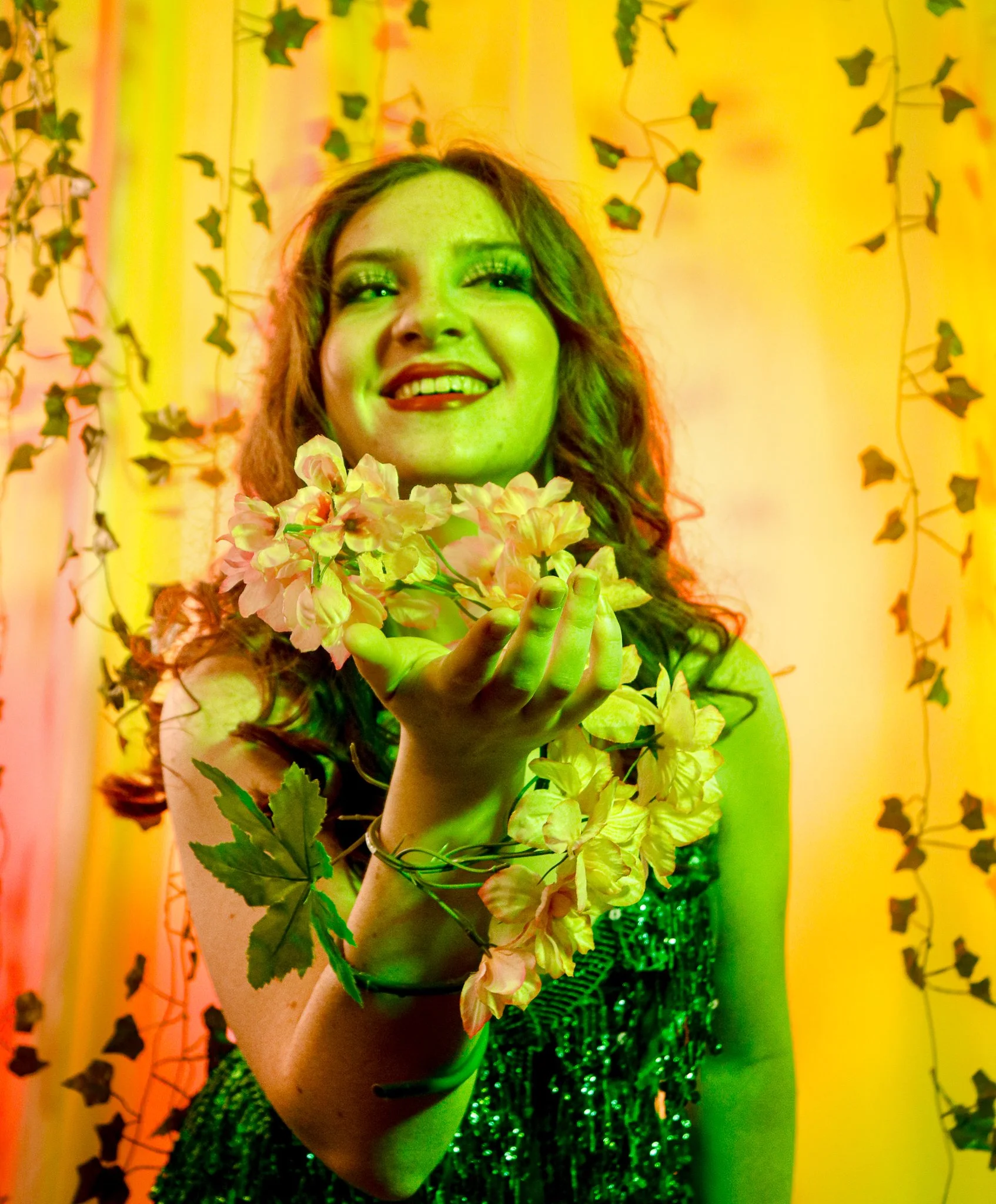A smiling woman with long wavy hair holds a bunch of yellow flowers in front of a backdrop decorated with ivy leaves and yellow lighting.