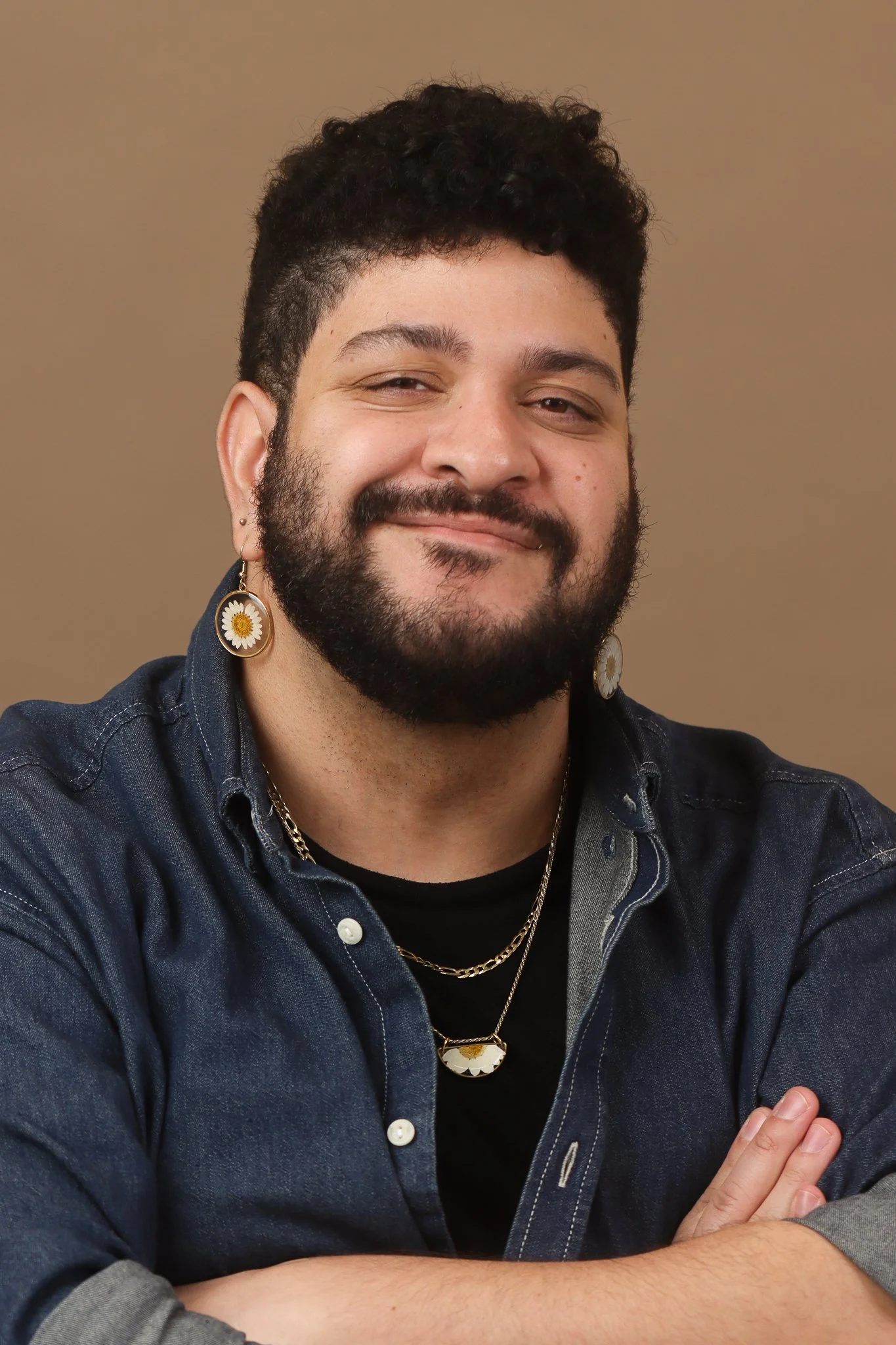 Close-up of a smiling young man with curly hair, beard, and earrings wearing a denim jacket and layered necklaces, against a neutral background.