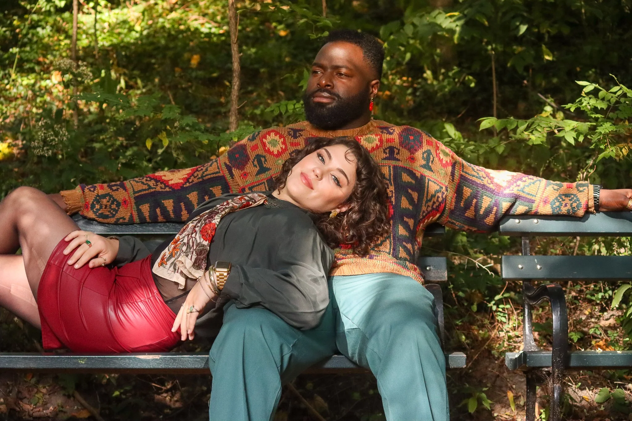Two people sitting on a park bench surrounded by green foliage. One person is lying on the other's lap, both looking relaxed.