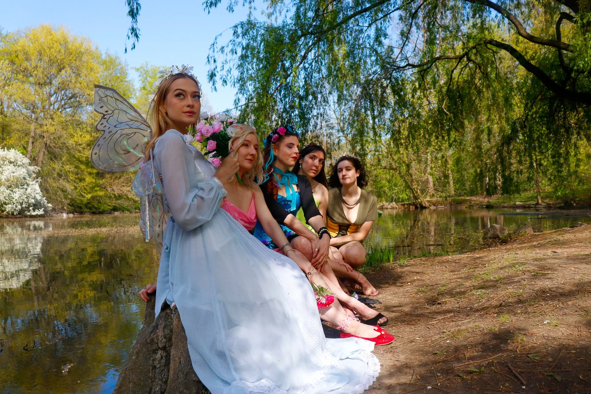 Group of women dressed as fairies, with wings and floral accessories, sitting by a lakeside in a forested area on a sunny day.