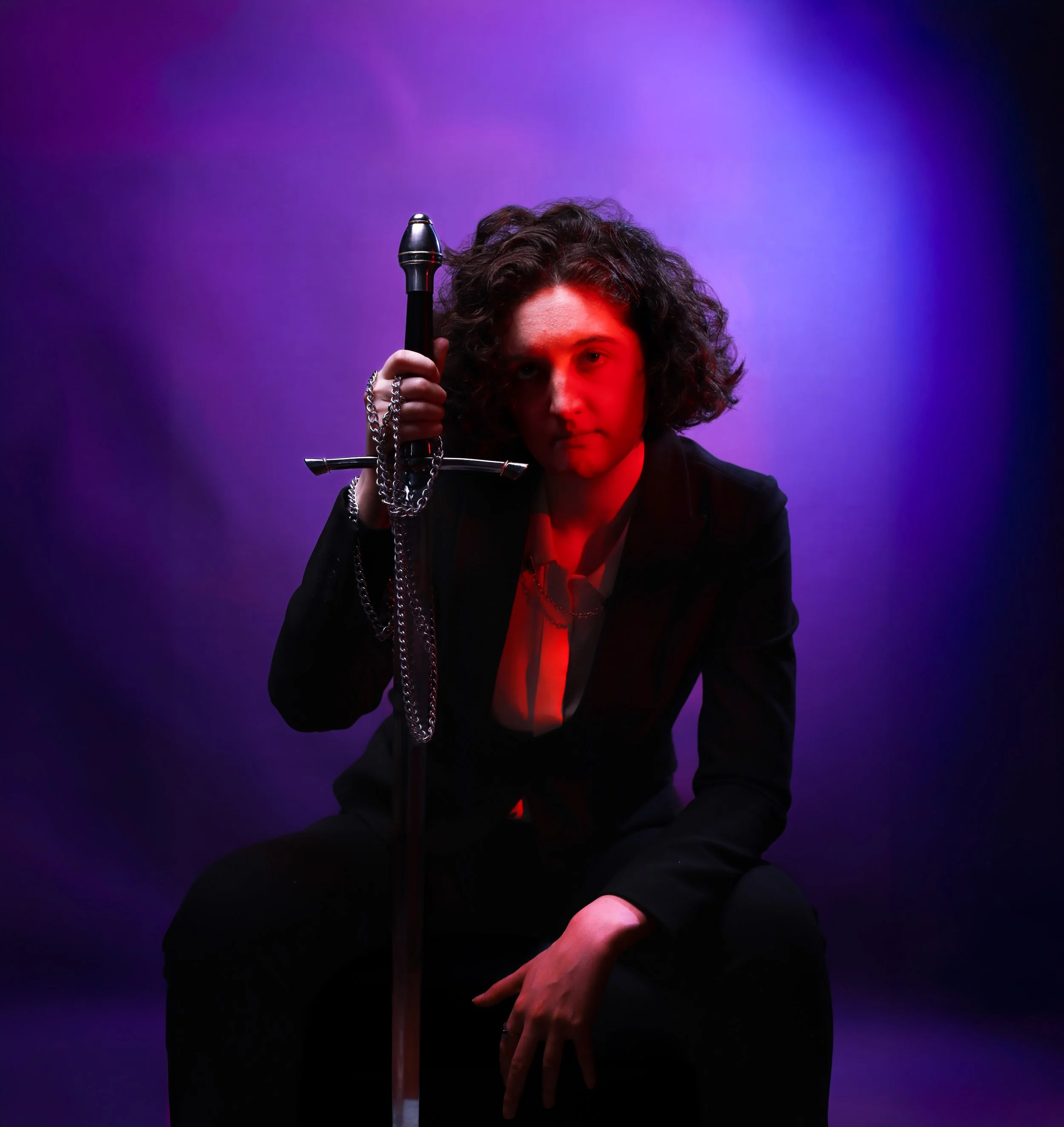 A person with curly hair holding a sword with a chain, illuminated by red and purple lighting.