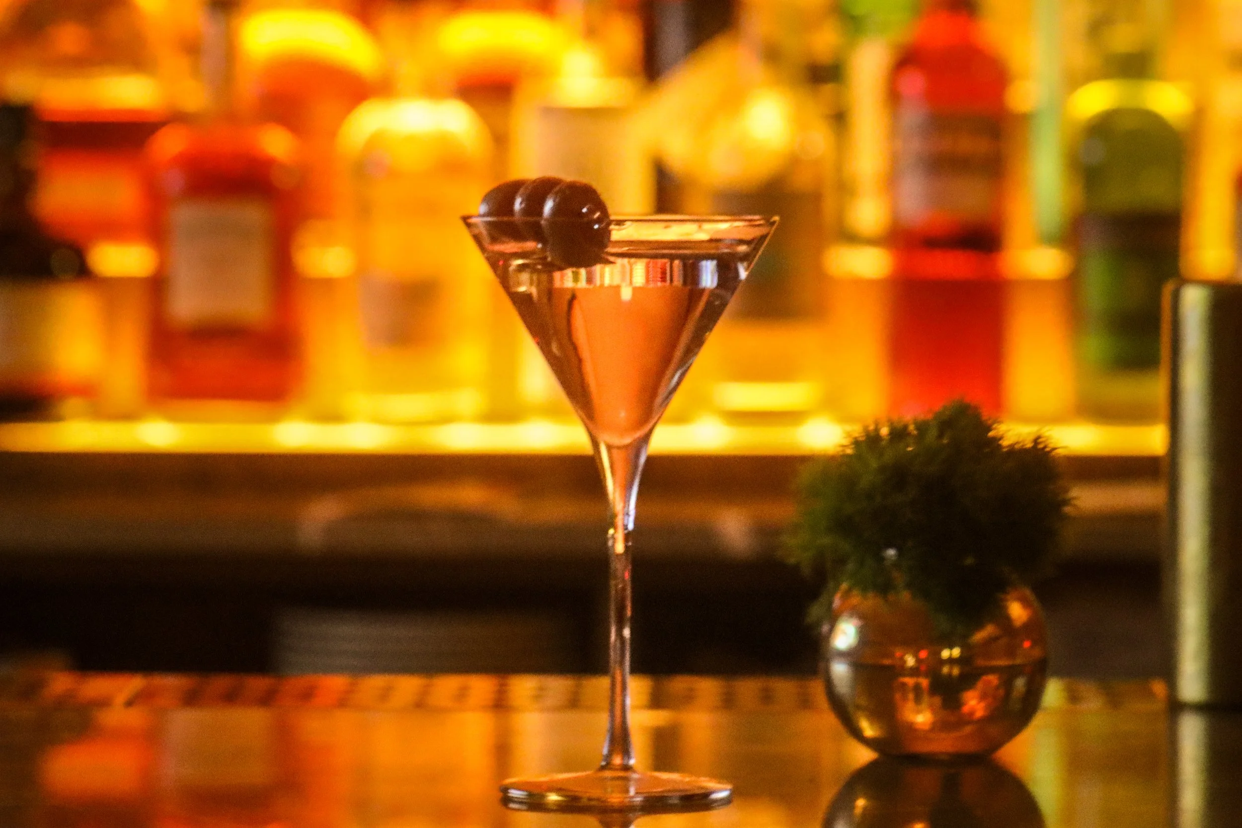 A cocktail glass with dark cherry garnishes sits on a bar counter with blurred colorful bottles in the background.